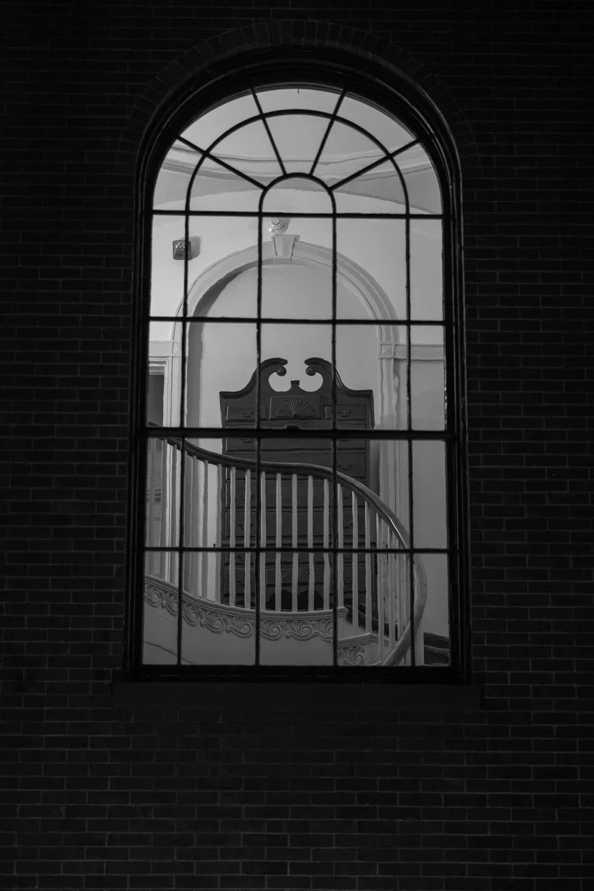 View through a large arched window showing a spiral staircase with ornate railing, an antique wooden cabinet, and decorative molding inside a building.