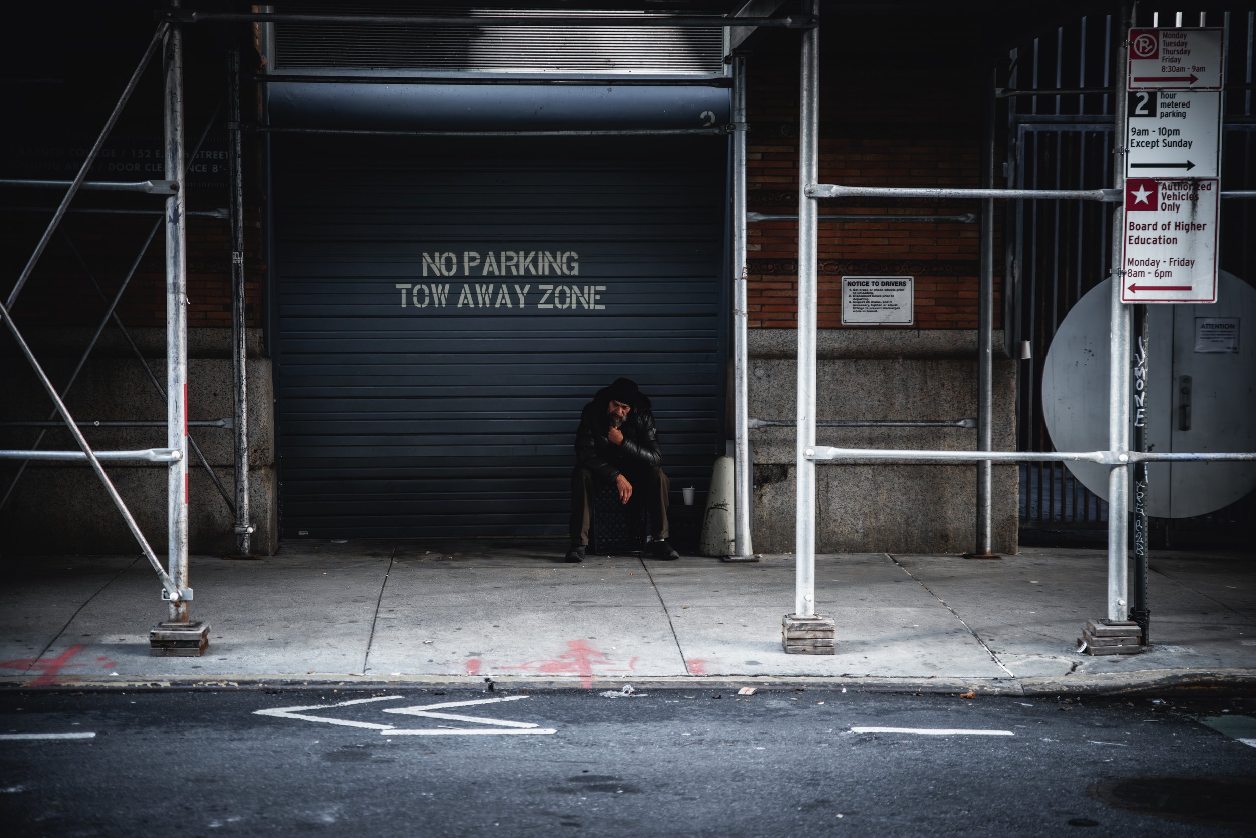 A man with long hair and a beard sitting on a crate under a roller shutter door, with a sign that says 'NO PARKING TOW AWAY ZONE' on the door. Surrounding scaffolding is present, and there are parking signs on the right side of the image.