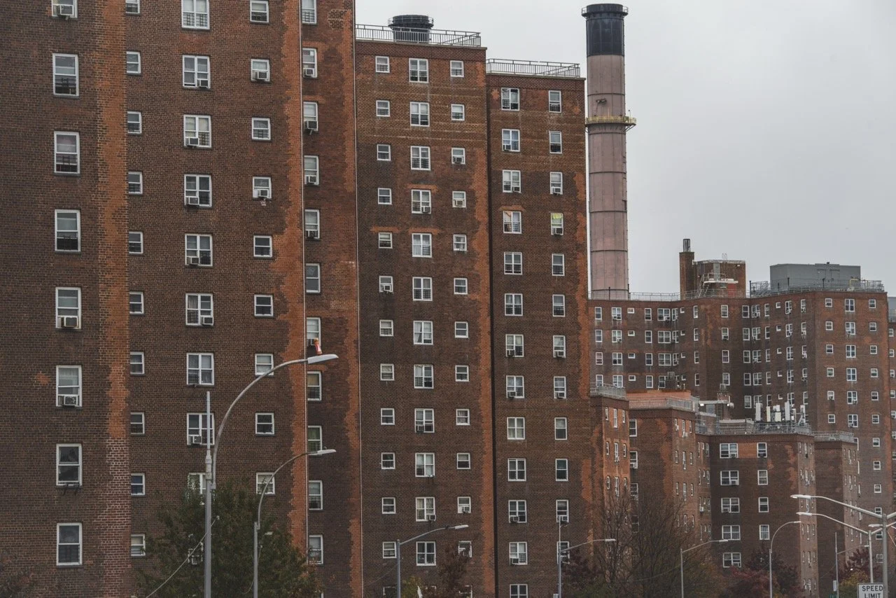 Multiple brick apartment buildings with numerous windows and a tall smokestack in an urban setting, overcast sky.
