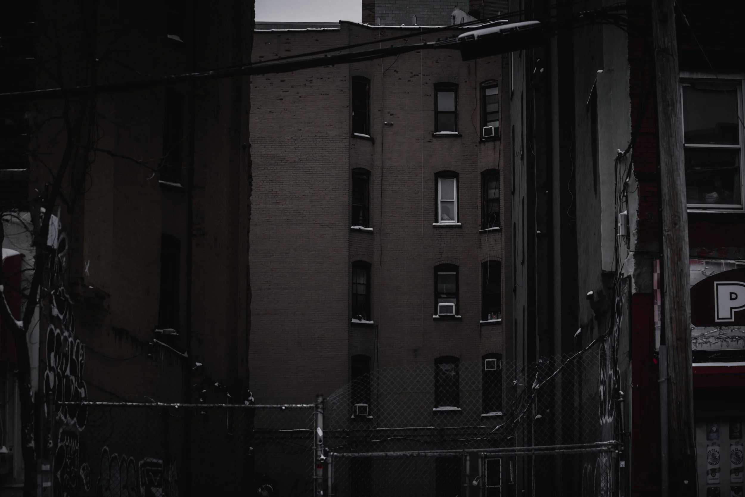 A view of a city alleyway with brick buildings, electrical wires, and a chain-link fence in the foreground, during winter with snow on the window ledges.