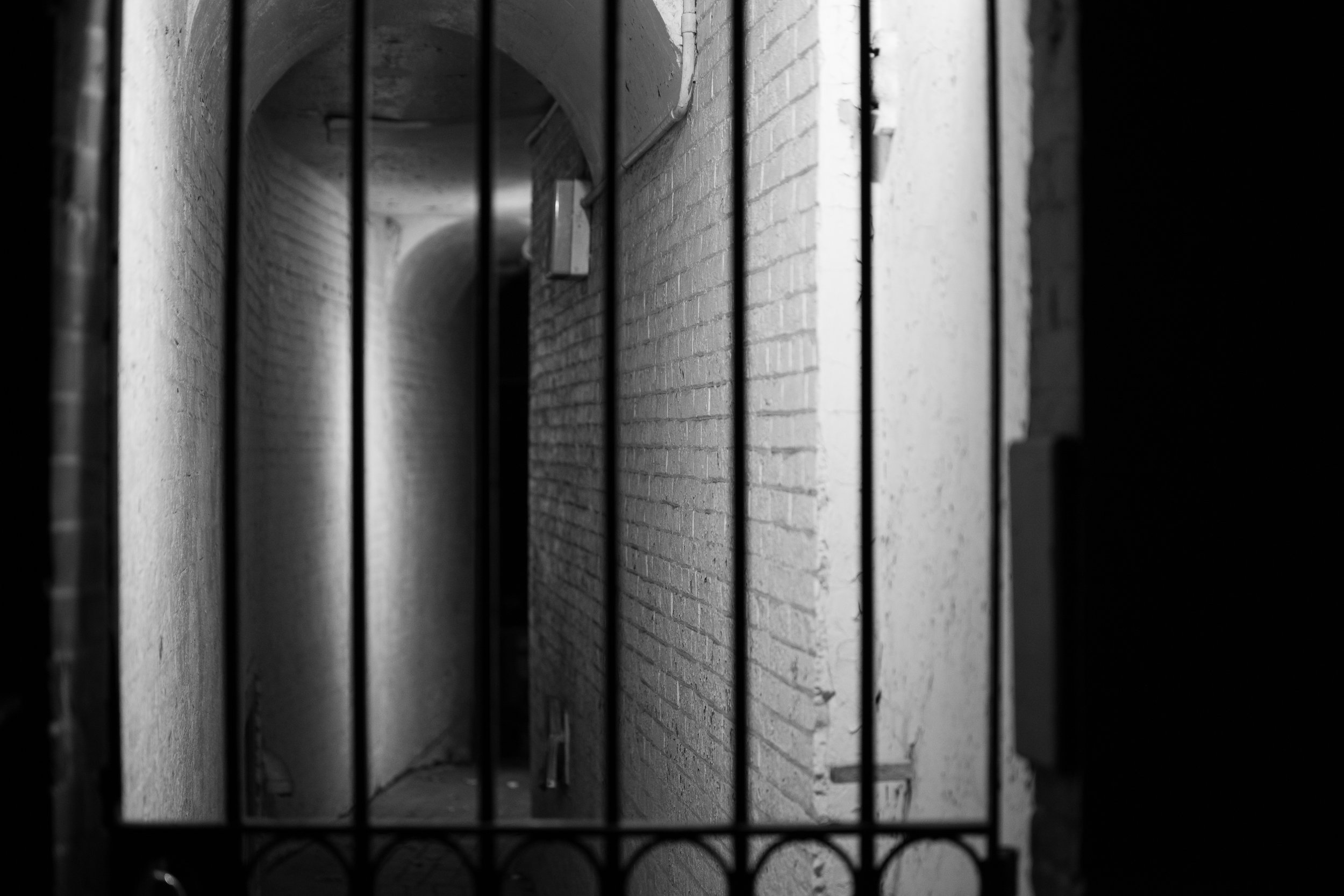 Black and white photo of an alleyway or corridor viewed through black metal bars, with a brick wall and an archway in the background.