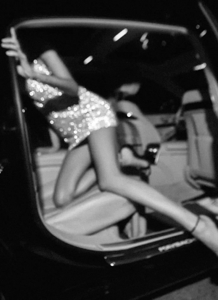 A woman with long hair wearing a sequined dress, sitting in a car, leaning against the door frame.