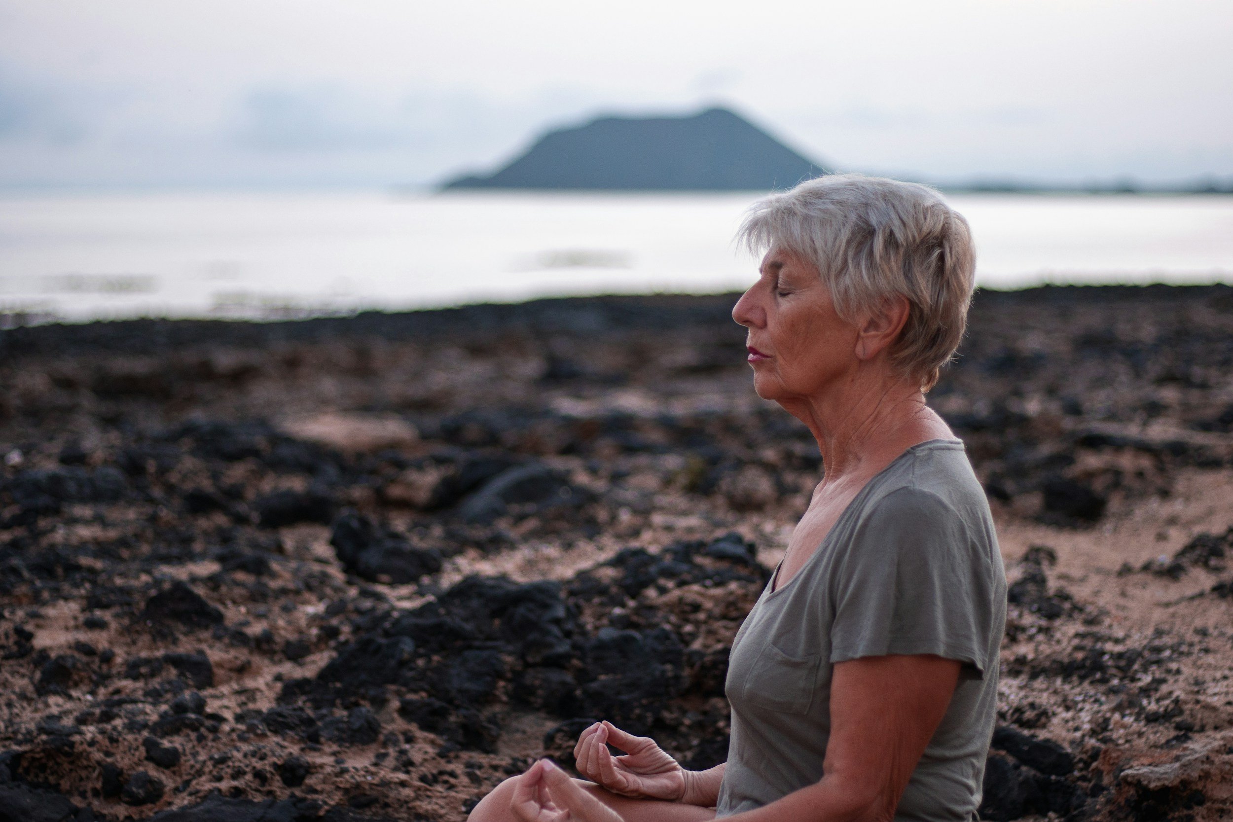 Una mujer mayor con cabello gris claro, meditando con los ojos cerrados en la playa de piedras, con un islote en el fondo y el cielo nublado.