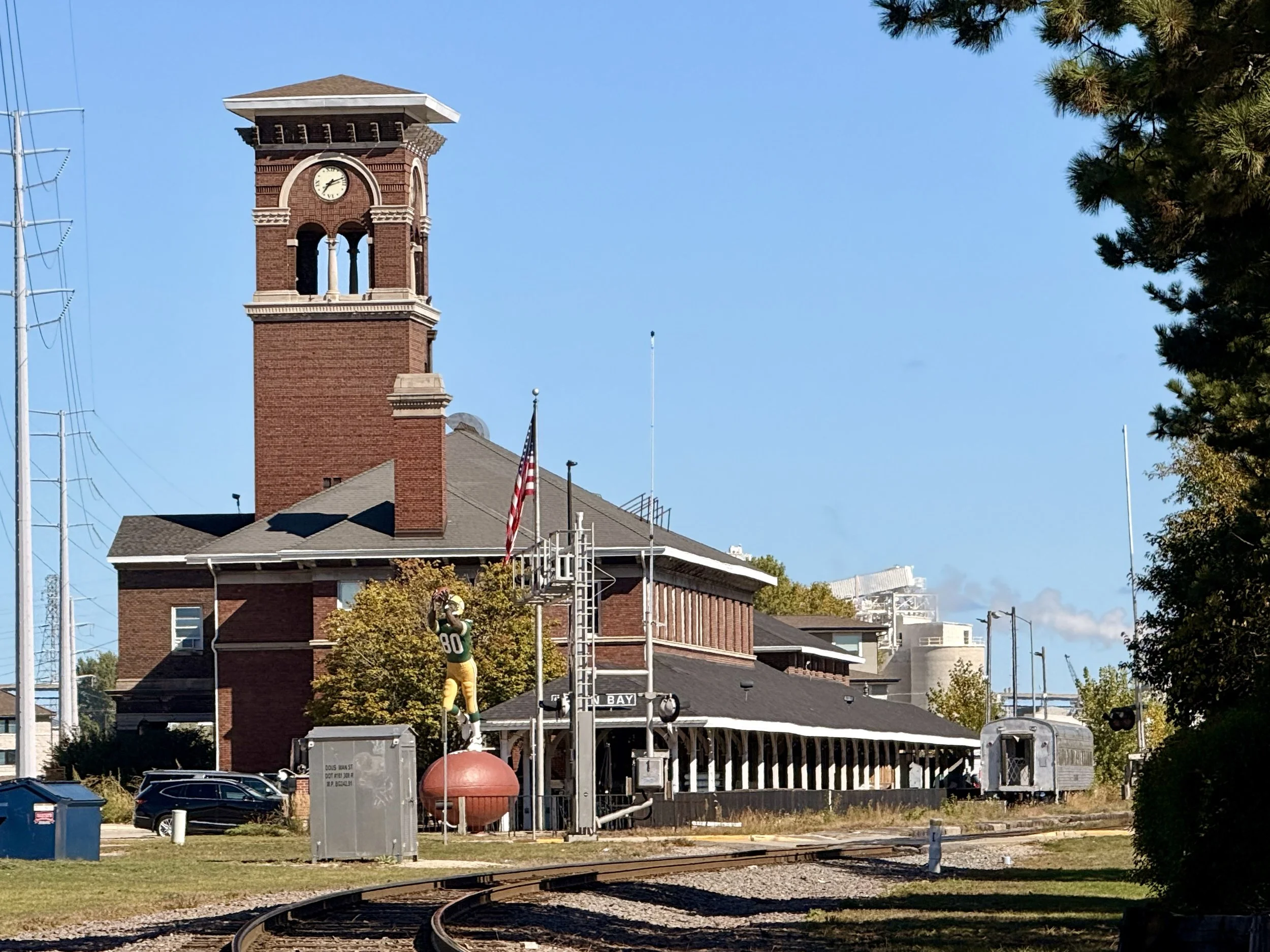 A brick clock tower with a white clock face and a small roof on top, situated behind a building with a porch. The scene includes overhead power lines on the left, an American flag, trees, and a train track in the foreground, with a clear blue sky ove