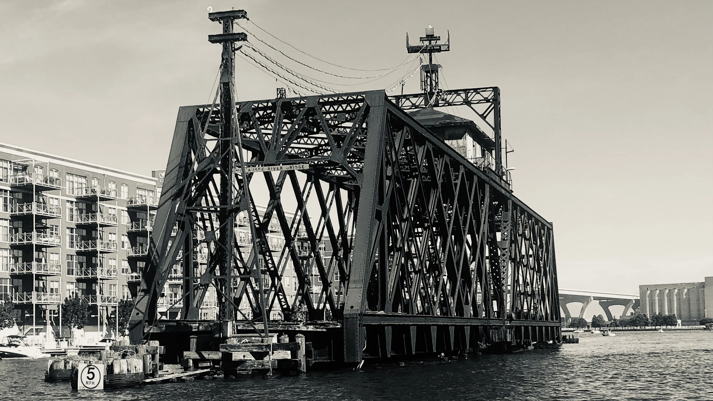 Steel bridge over a waterway with a nearby apartment building and highway in the background, black and white photo.