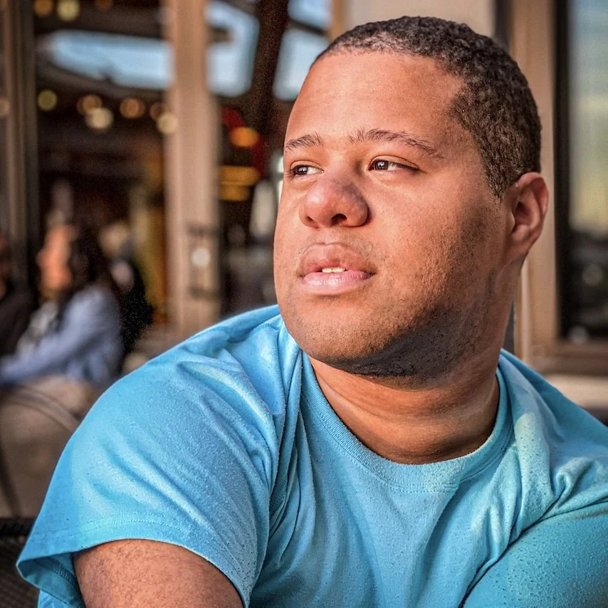 A young man with short curly hair, wearing a bright blue shirt, looking off into the distance at a cafe or restaurant with out-of-focus people and warm lighting in the background.