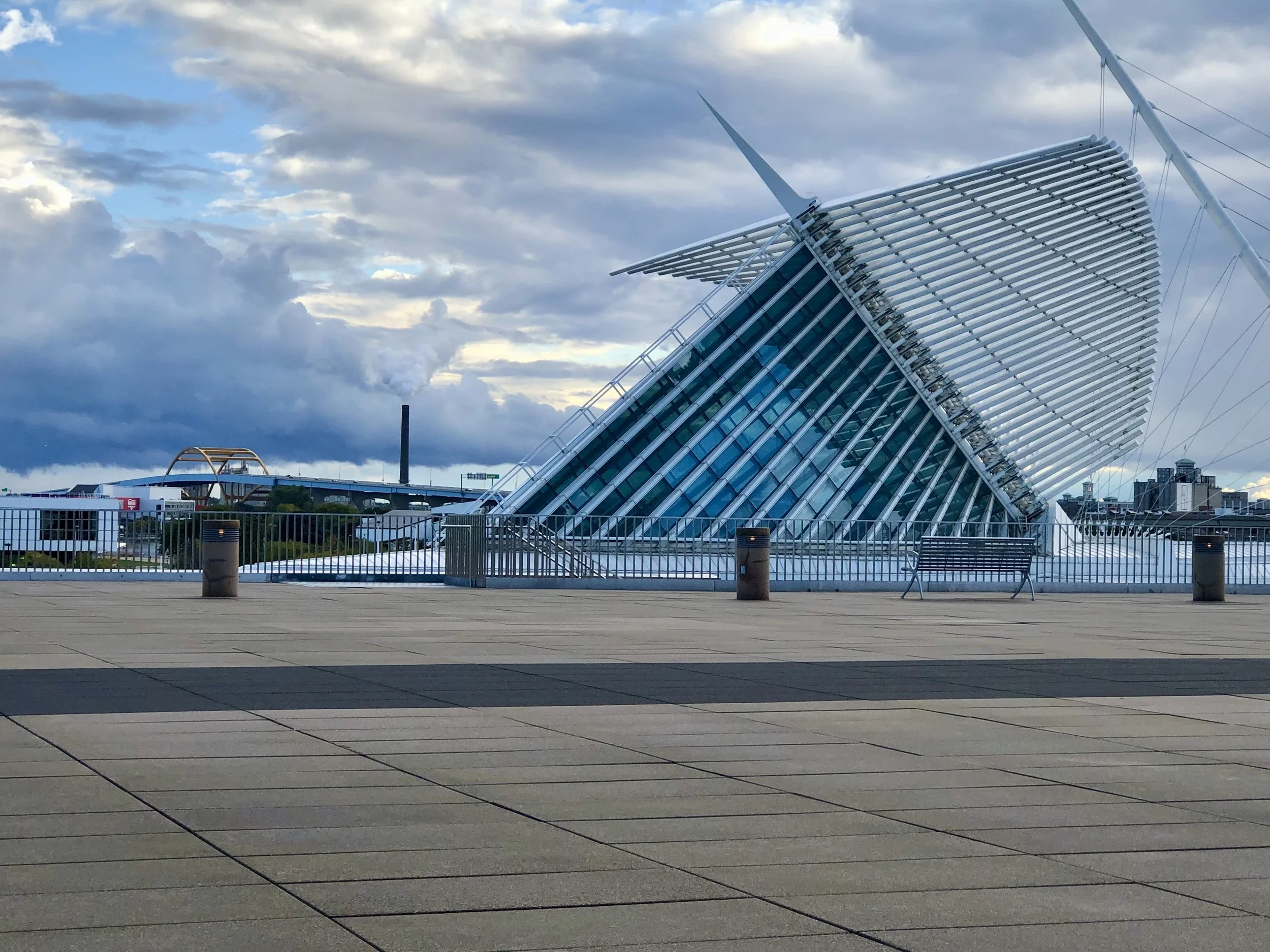 A modern building with a unique architectural design featuring a large, curved, white, ribbed structure with sharp, pointed spires. The building is surrounded by an open, paved area with a fence and lamps, under a cloudy sky.