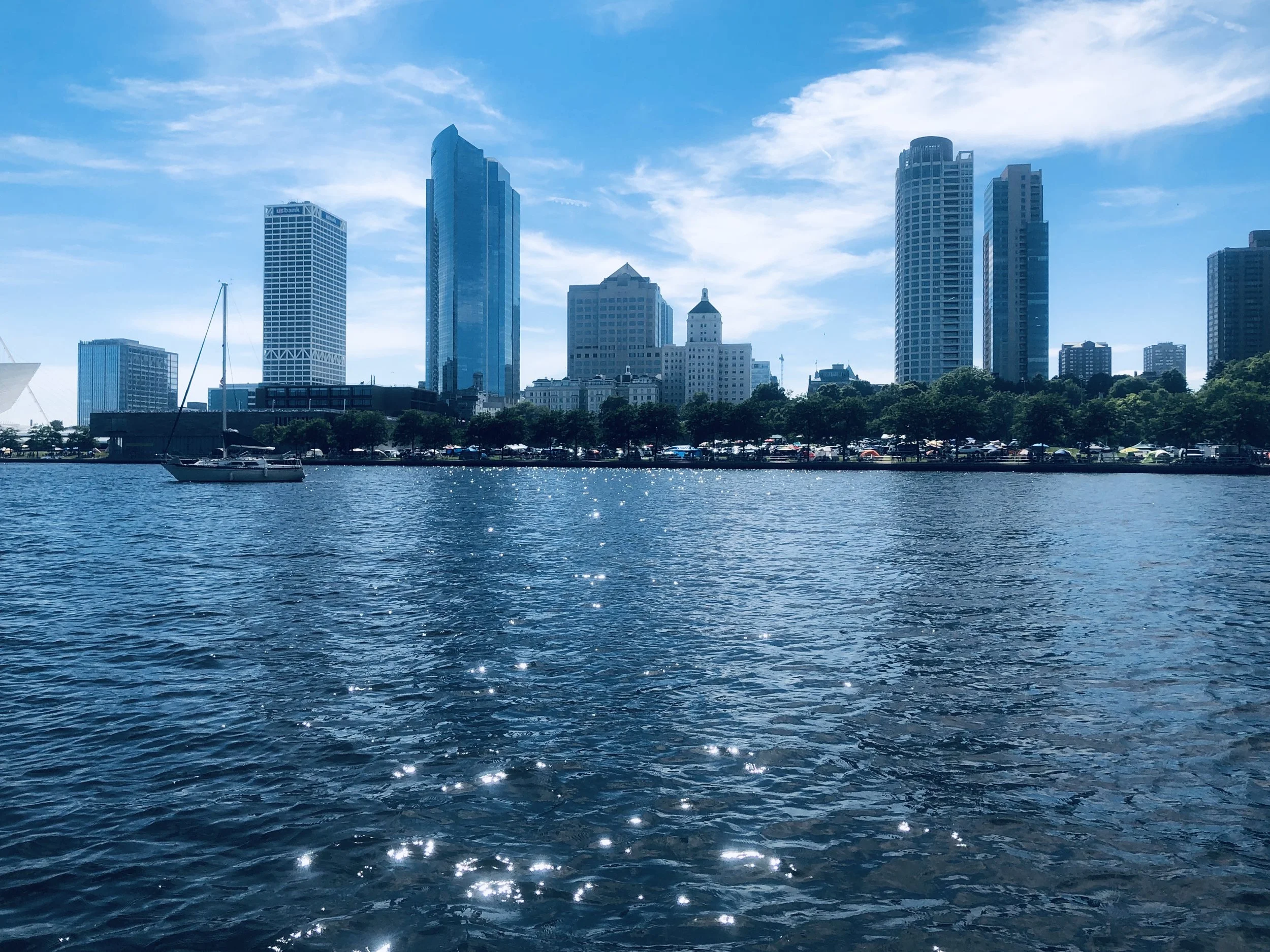 View of a city skyline with modern skyscrapers across a body of water, with a sailboat in the foreground and a partly cloudy blue sky.