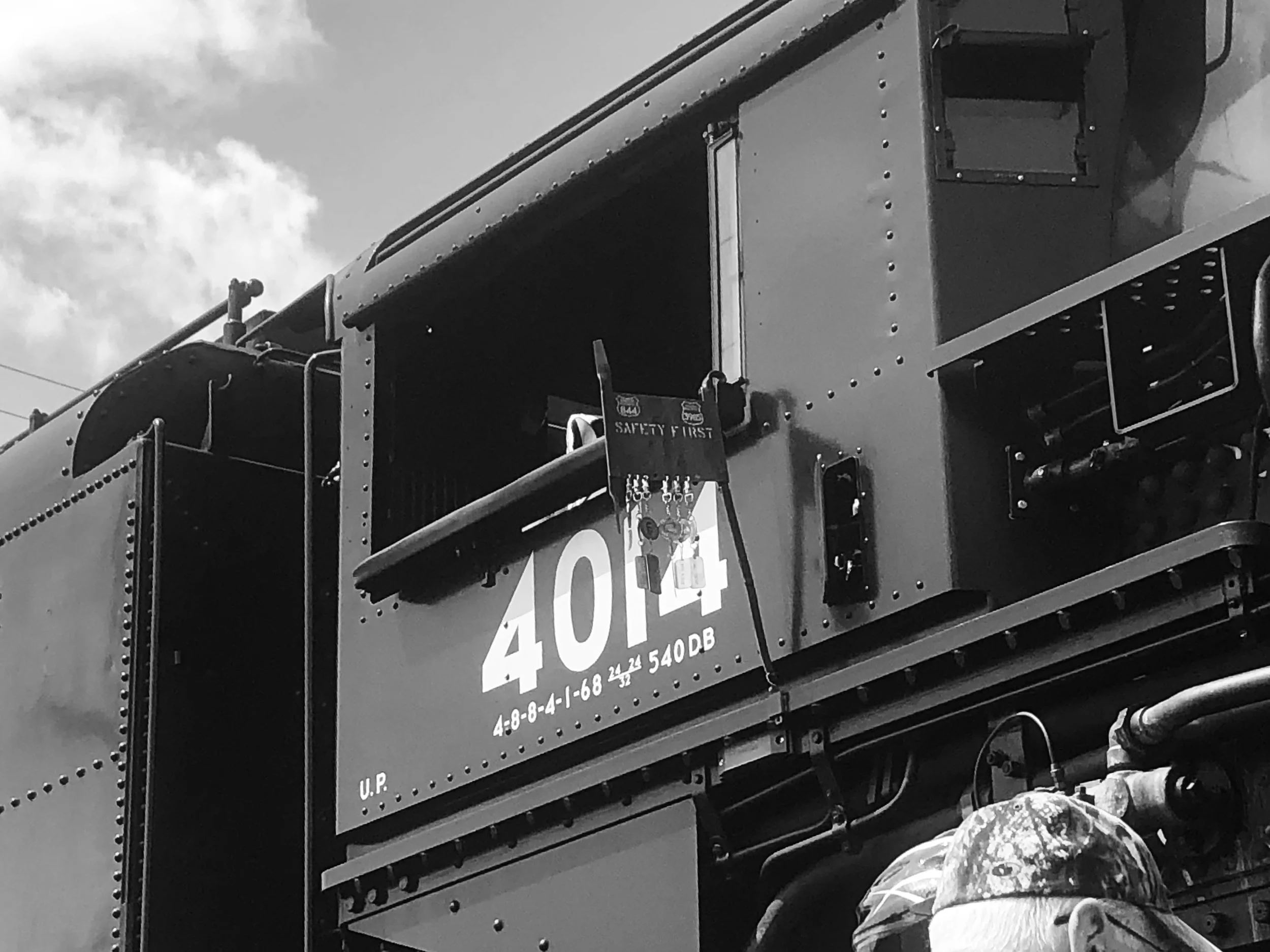 Close-up of a steam locomotive with a sign indicating safety precautions and other markings, with a partly cloudy sky in the background.