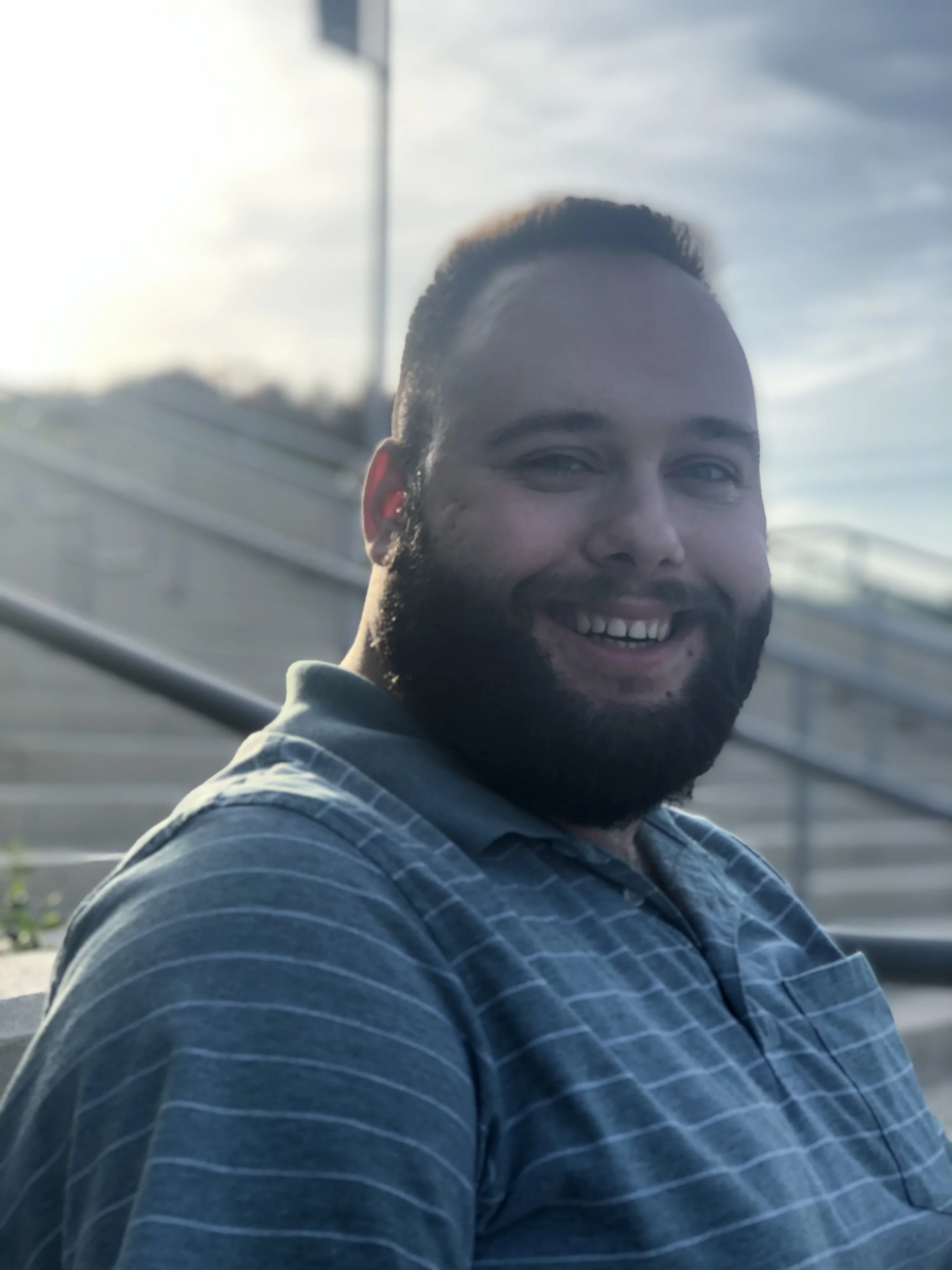 A smiling man with short dark hair and a beard, wearing a striped shirt, sitting outdoors during daytime with a building and cloudy sky in the background.