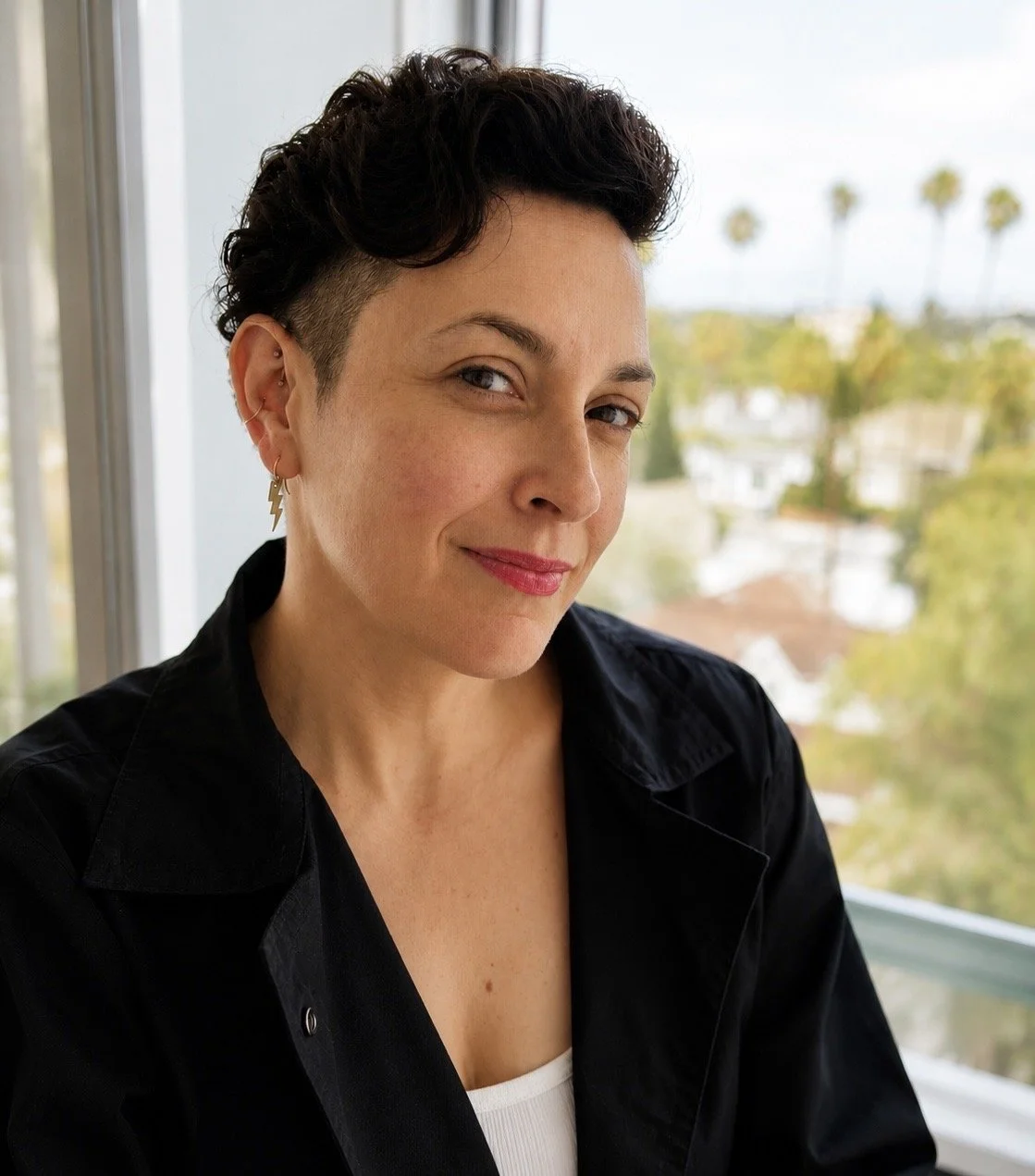 A woman with short dark wavy hair, wearing earrings with lightning bolt designs, smiling with a sidelong glance, sitting in front of a window with trees and palms outside.
