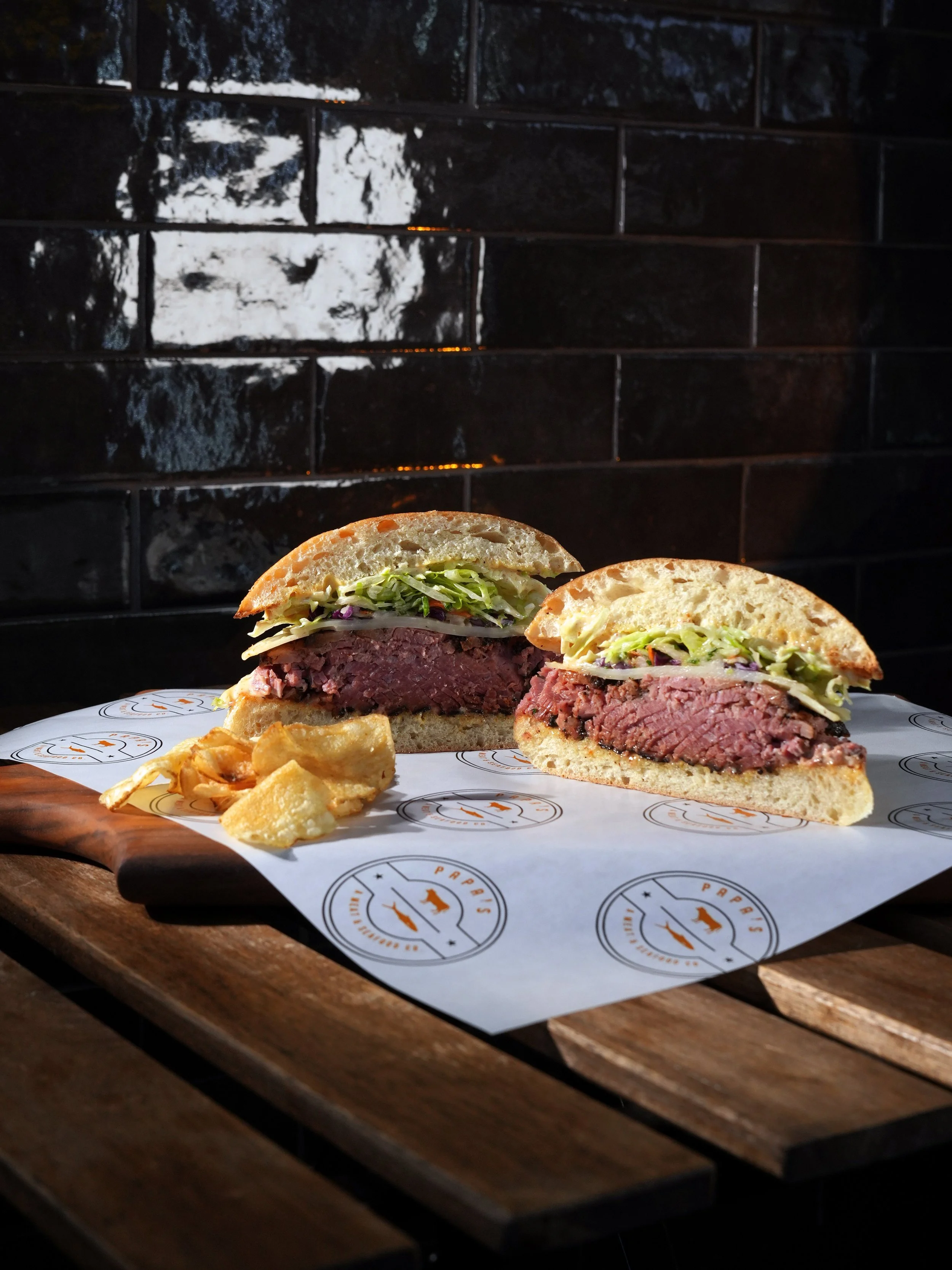 A sliced American-style burger with a beef patty, lettuce, onion, and bun, served on parchment paper with fries on a wooden tray in front of a black tiled wall.