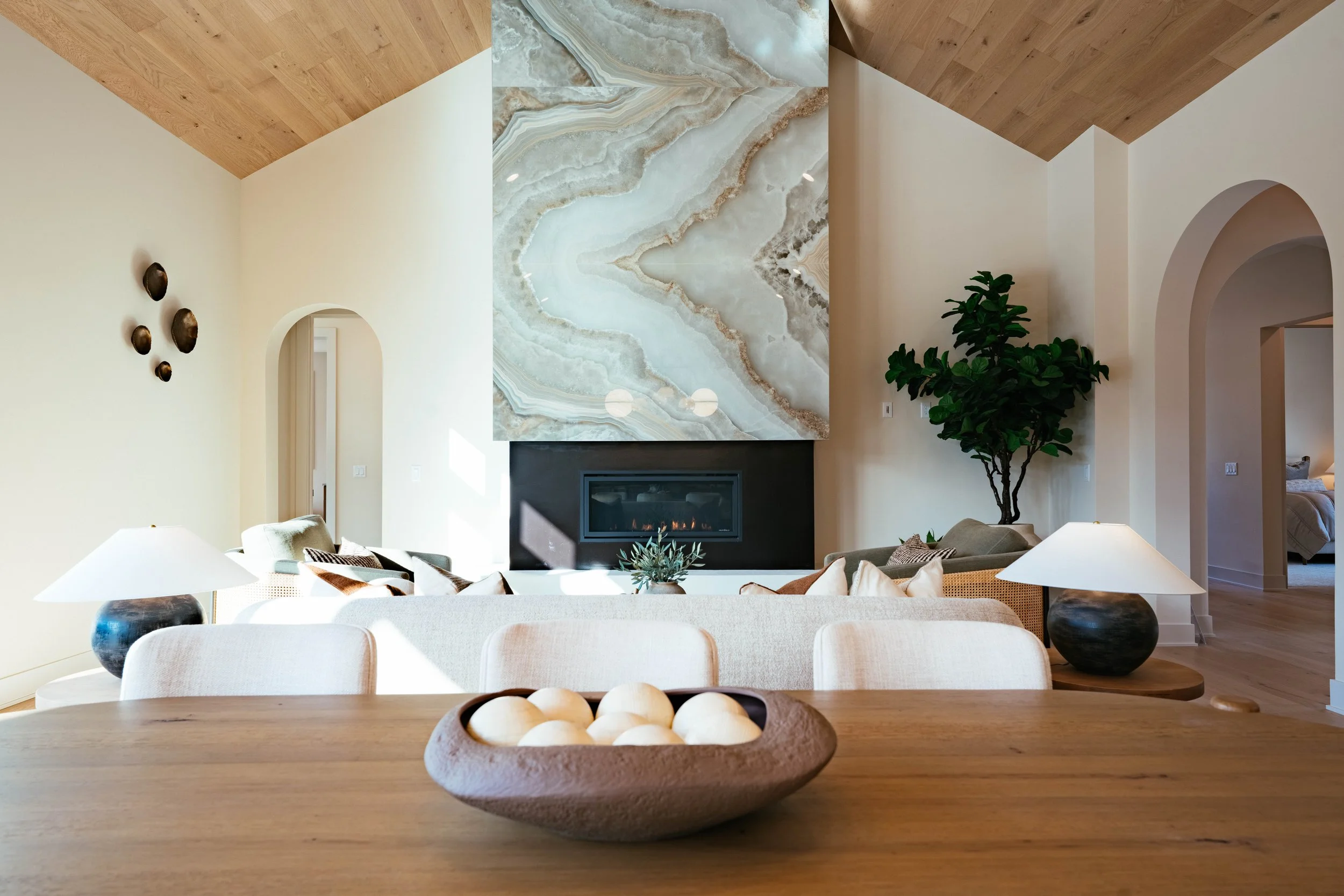 A living room with a large marble fireplace, a wooden ceiling, beige walls, a houseplant, and multiple beige chairs and white lamps.