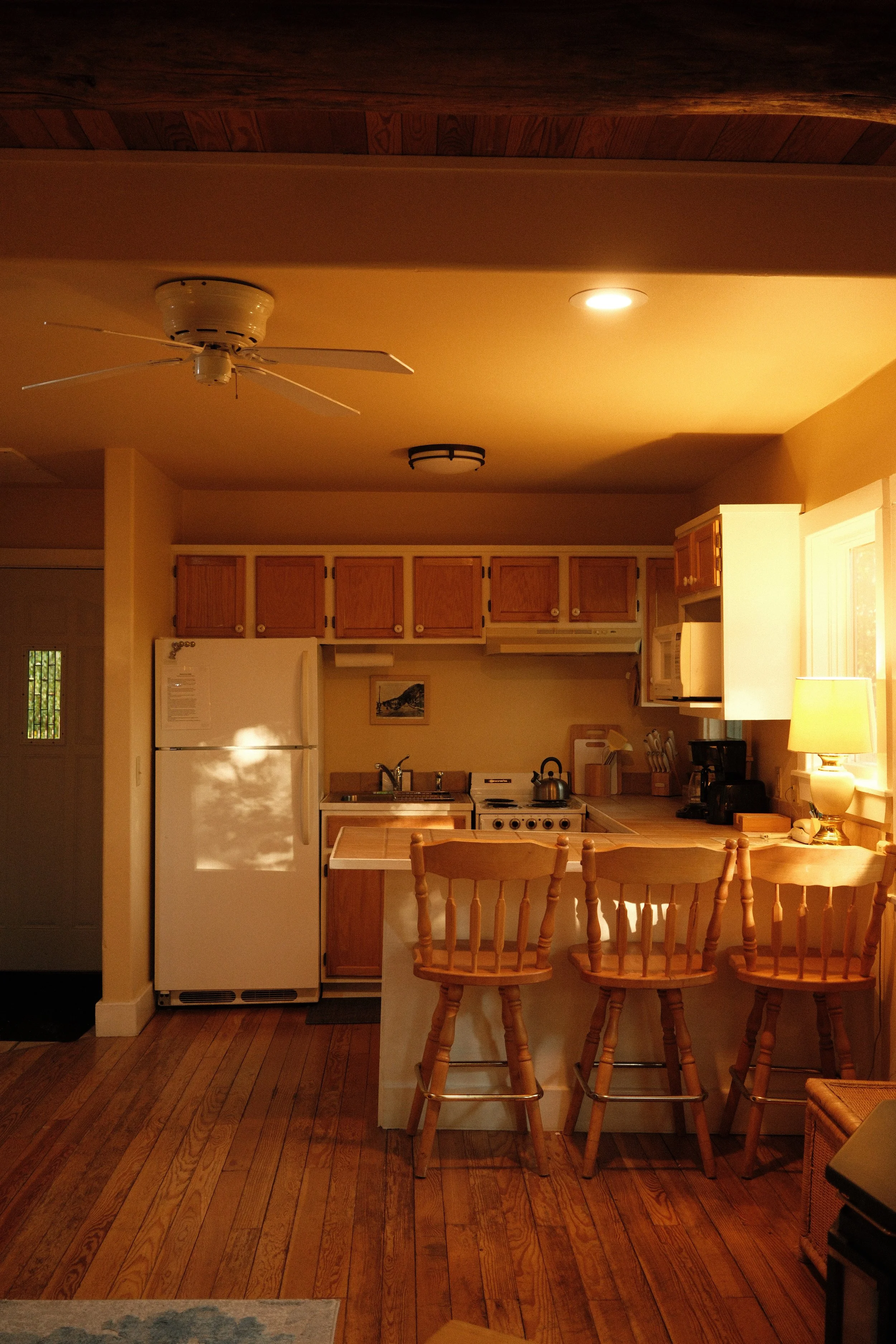 A cozy kitchen with wooden cabinets, white refrigerator, stovetop with kettle, and a breakfast bar with wooden chairs. Warm lighting and hardwood flooring create a inviting atmosphere.