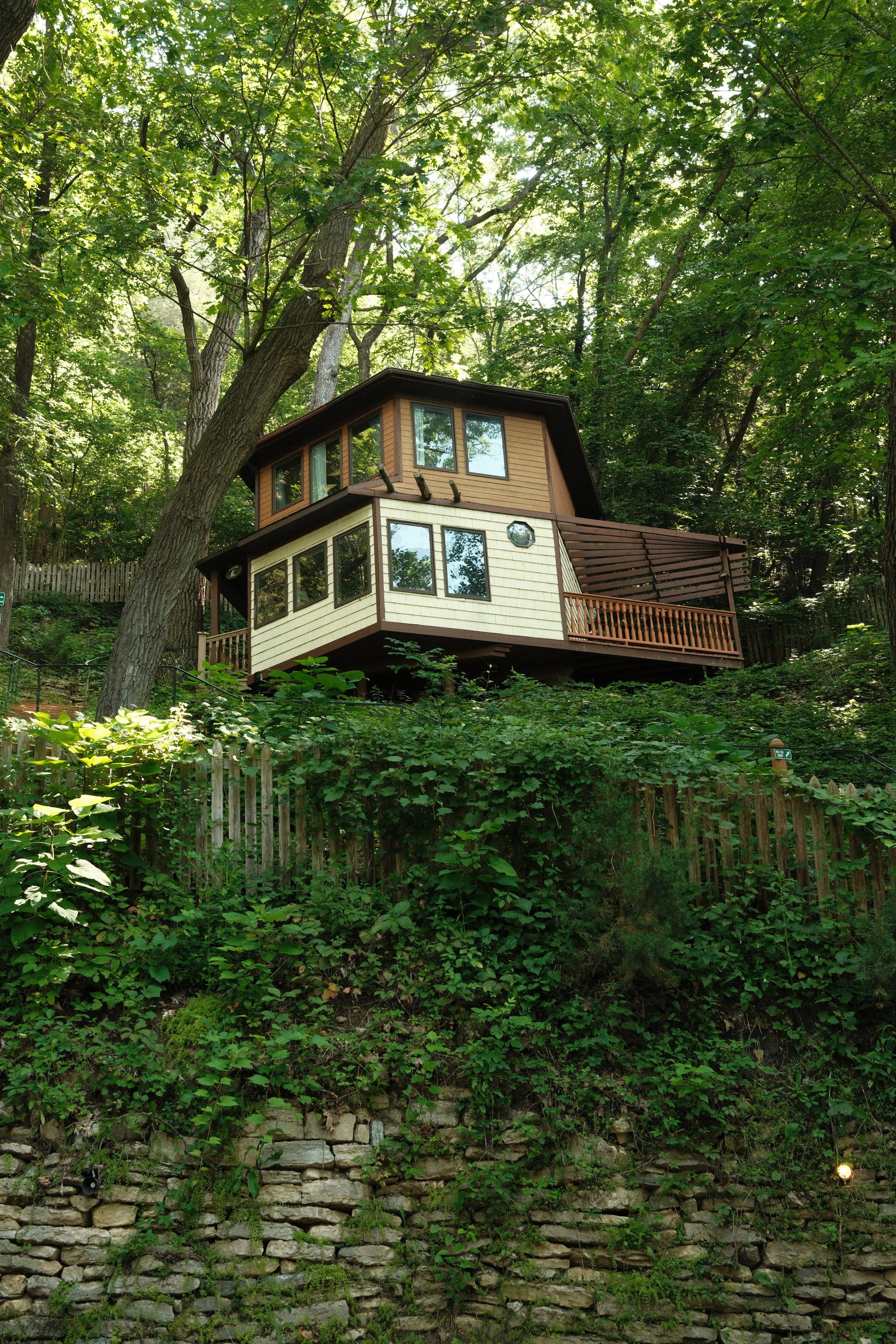 A treehouse perched on a hillside surrounded by dense green trees and foliage, with large windows and a wooden balcony.