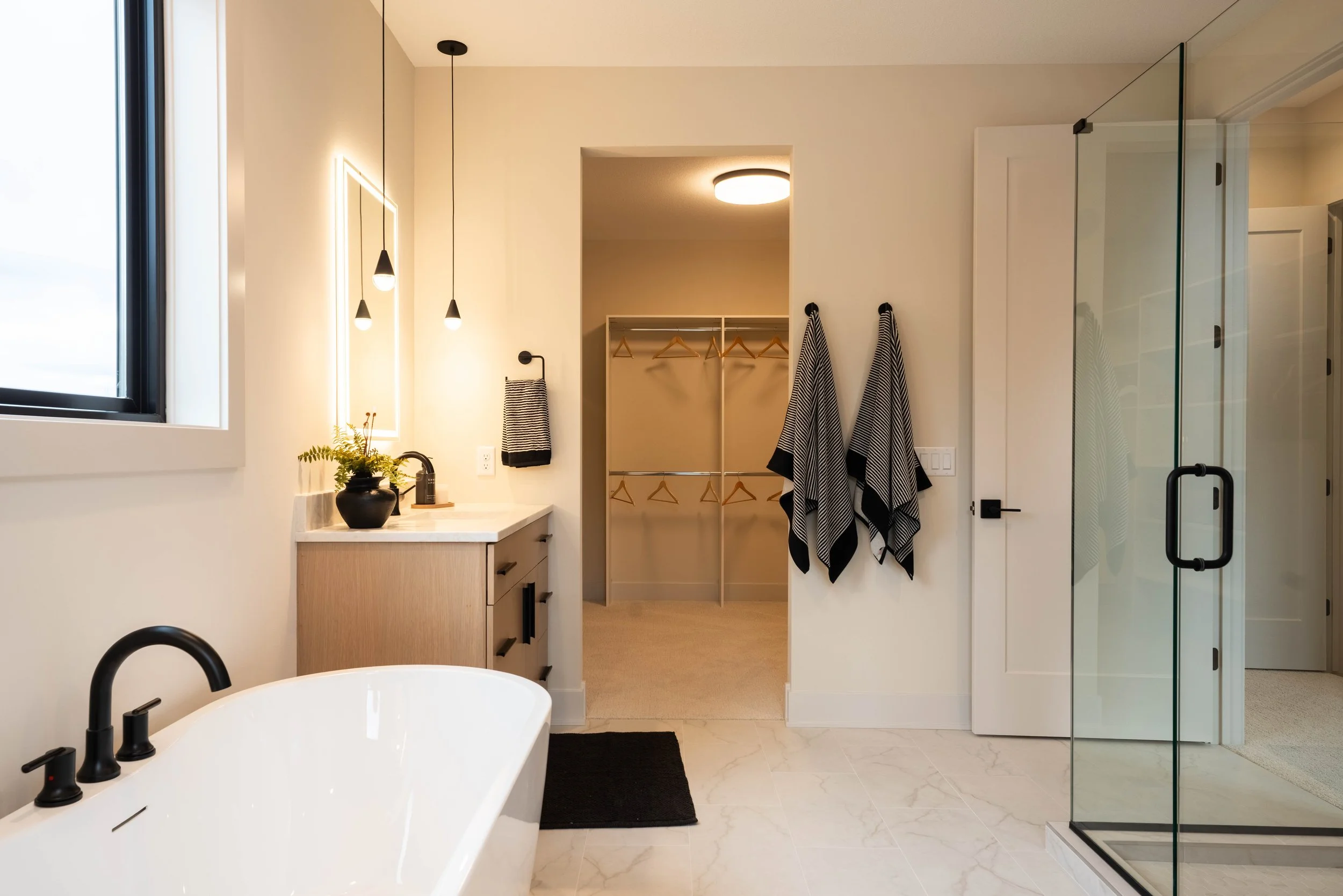 Modern bathroom with a freestanding bathtub, black fixtures, a window, a vanity with a plant, hanging towels, and a walk-in closet with wooden hangers, a ceiling light, and a glass shower door.