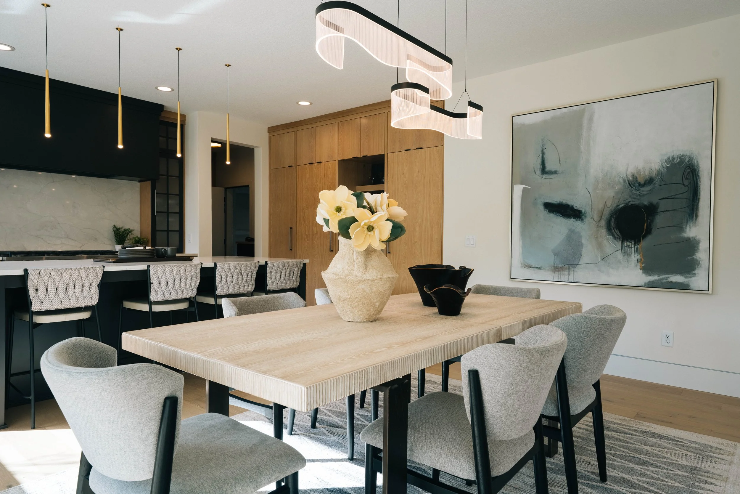 Modern dining and kitchen area with a light wood table, gray upholstered chairs, a decorative vase with white flowers, abstract wall art, black and gold pendant lights, and wooden cabinetry.
