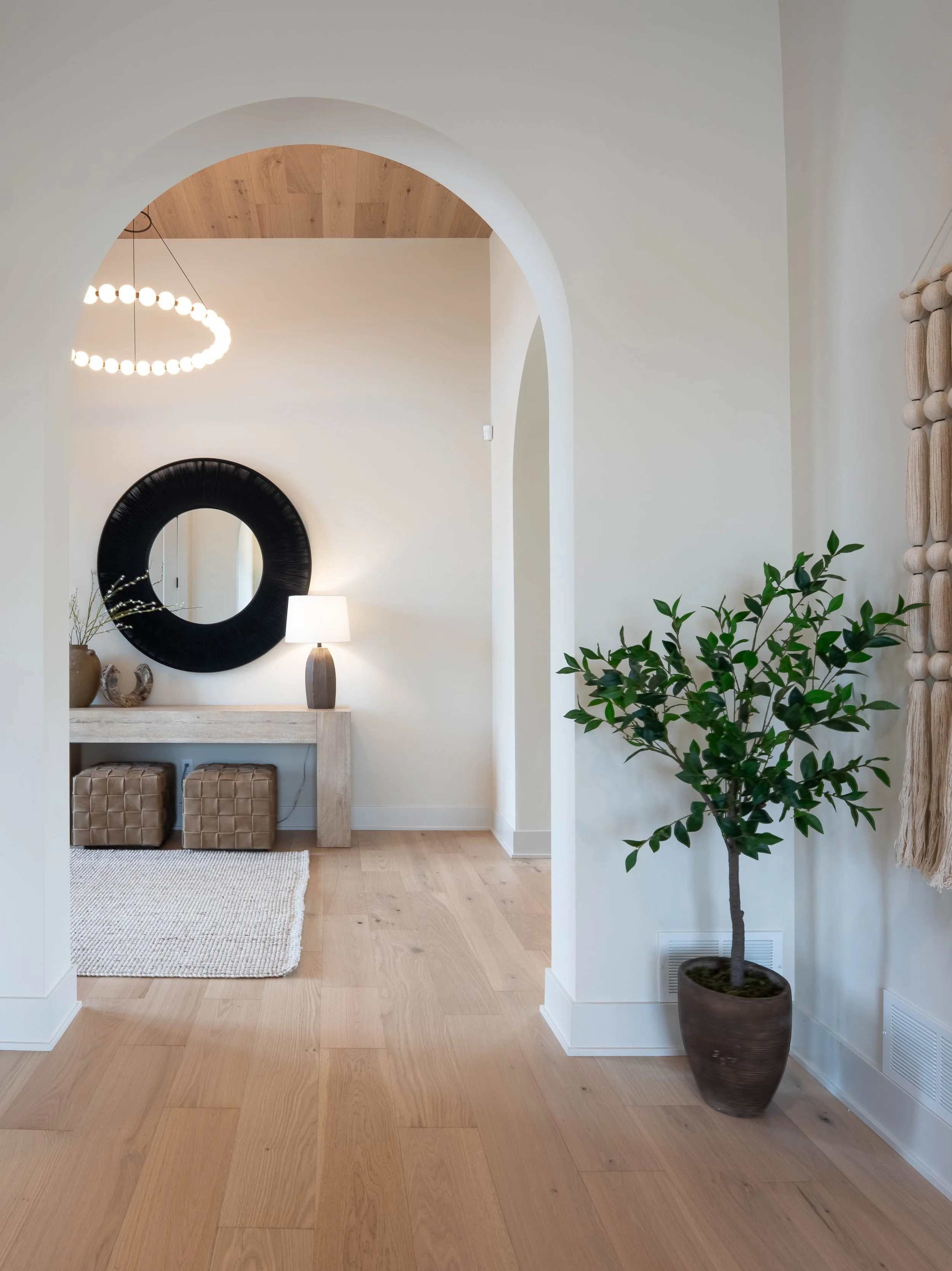 A minimalist living space with a large potted tree on the right, a console table with decorative vases and a lamp, a circular black mirror above the table, and a beige armchair with a woven ottoman underneath. The hardwood floor is light-colored and 