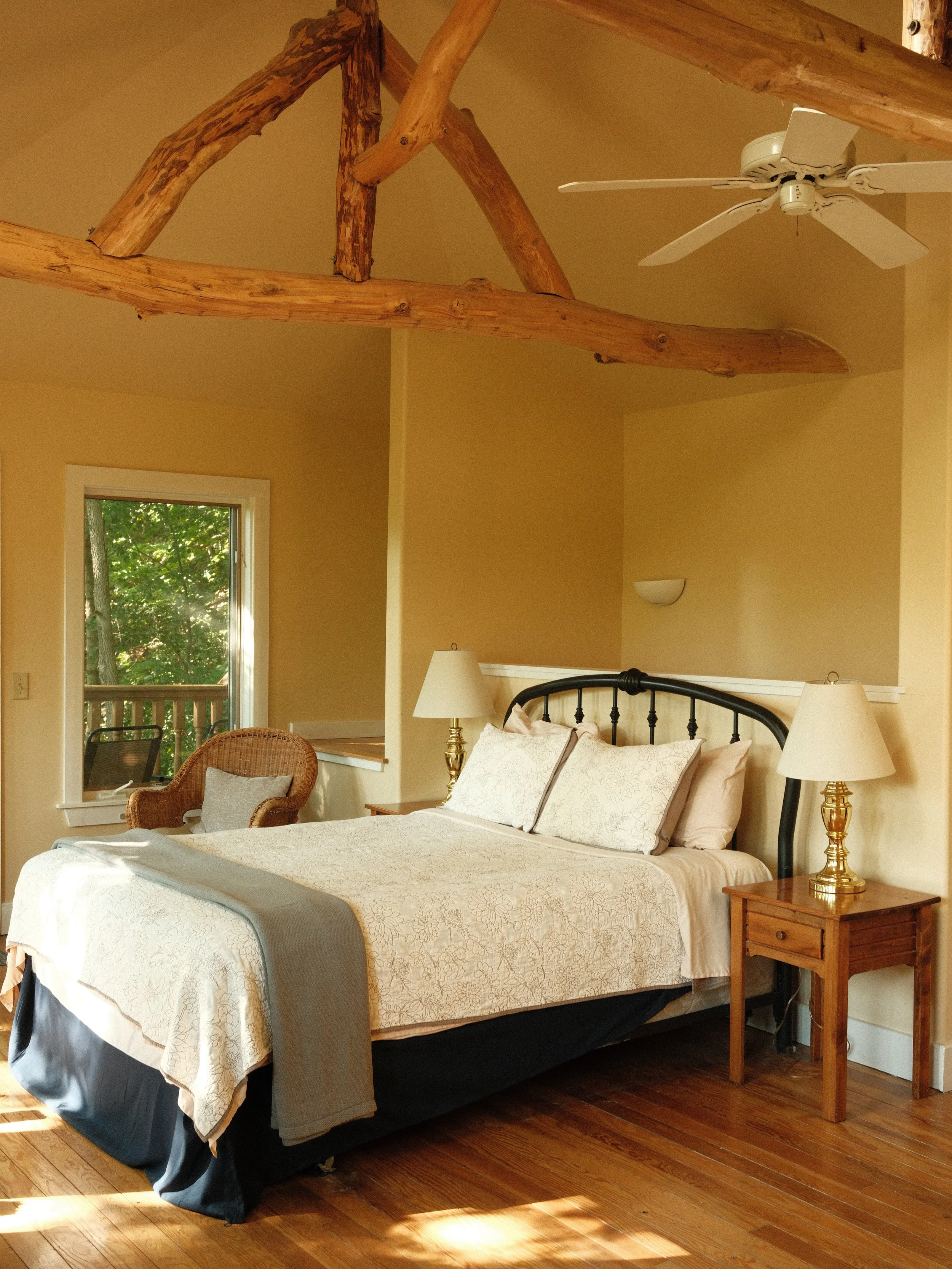 A bedroom with a bed, nightstand, lamp, wicker chair, window showing green trees outside, and a wooden ceiling with exposed beams and a ceiling fan.