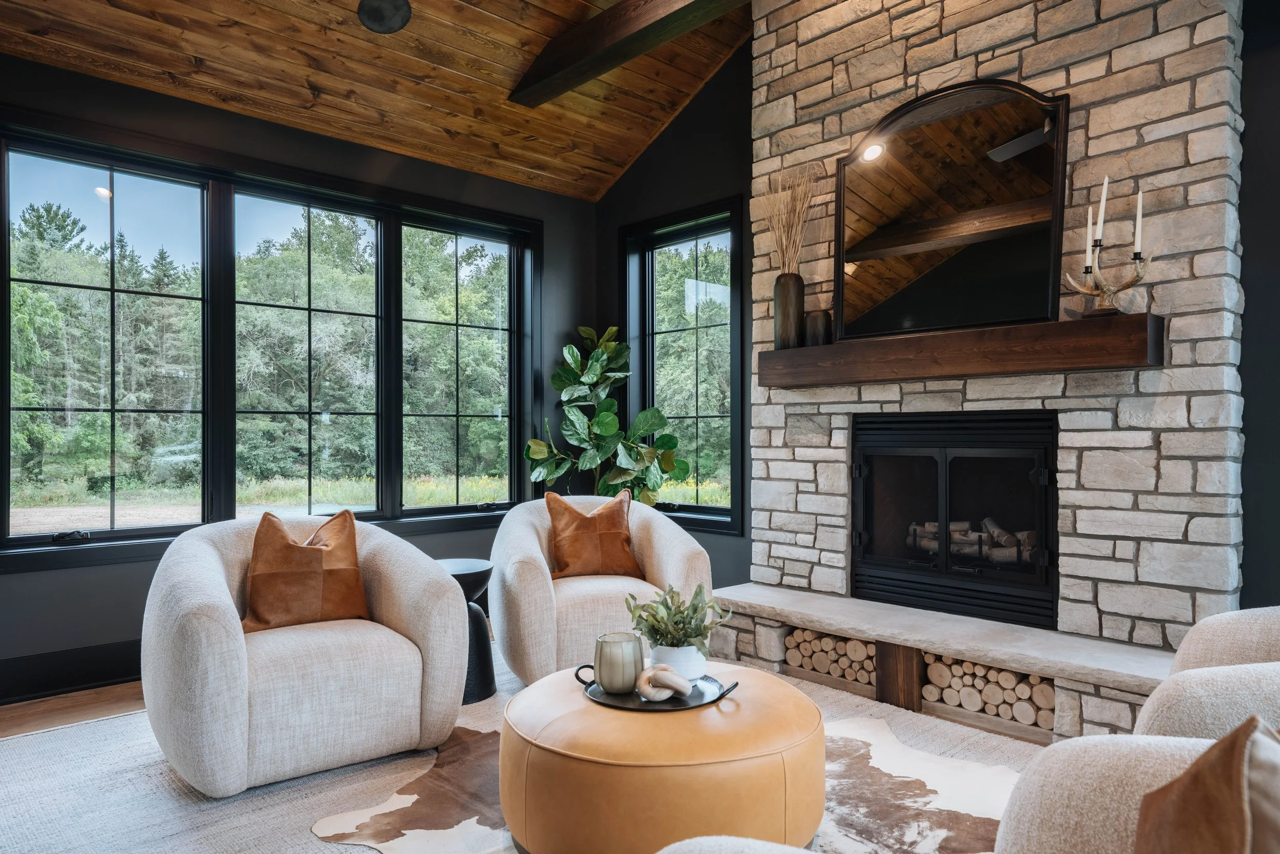 Living room with large windows showing green trees, a stone fireplace with wood logs underneath, a mirror above the fireplace, a potted plant, and beige upholstered chairs with brown pillows around a small round table with decorative items.