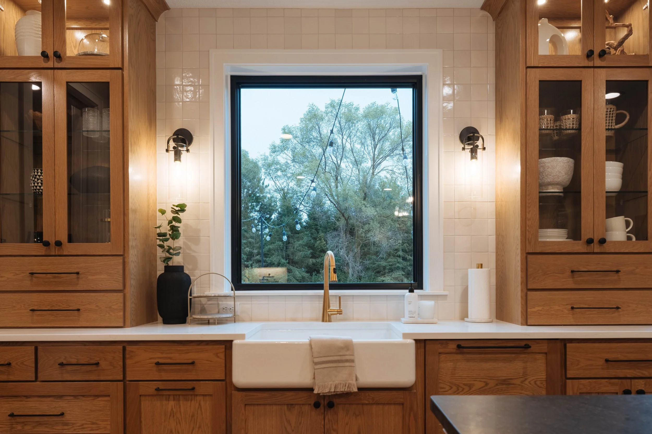 Kitchen with wooden cabinets and a large window showing trees outside. There is a white farmhouse sink with a gold faucet, a black vase with a plant, and a dish towel hanging on the sink. The cabinets have glass doors displaying dishes and glassware.