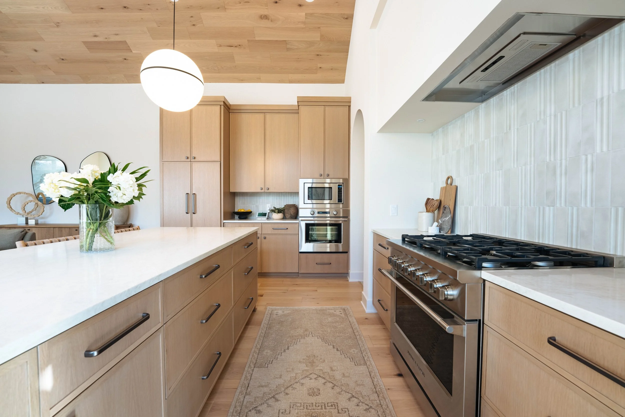 A modern kitchen with light wood cabinets, a white island with a vase of white flowers, stainless steel appliances including a microwave and oven, a gas stove, and a light-colored tile backsplash. There is a pendant light hanging from a wood-paneled ceiling, a beige area rug on the wooden floor, and decorative items near the stove.