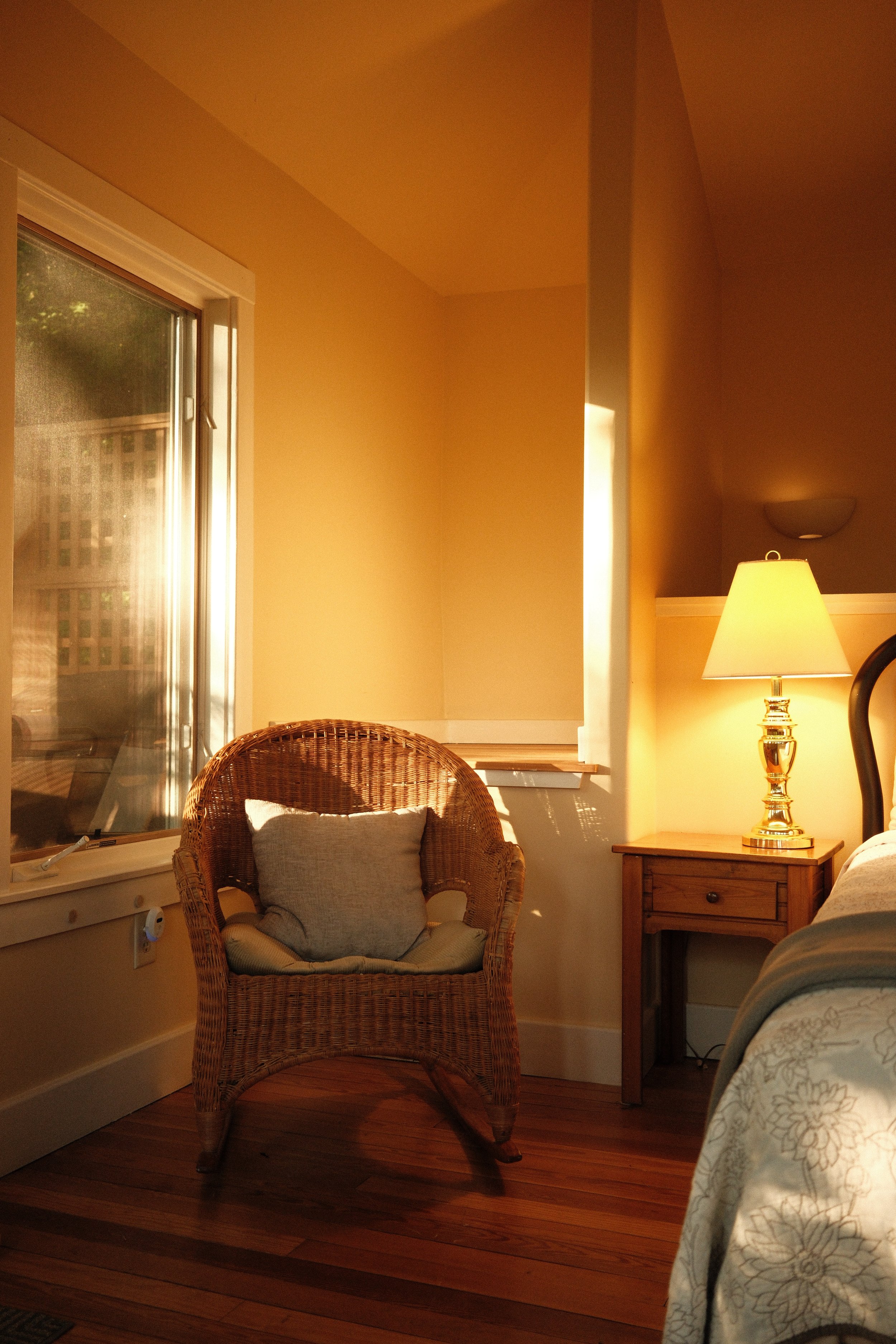 A cozy bedroom corner with a wicker armchair with a pillow, a wooden nightstand with a lamp, and a window with closed blinds letting in warm sunlight, featuring hardwood flooring and yellow painted walls.