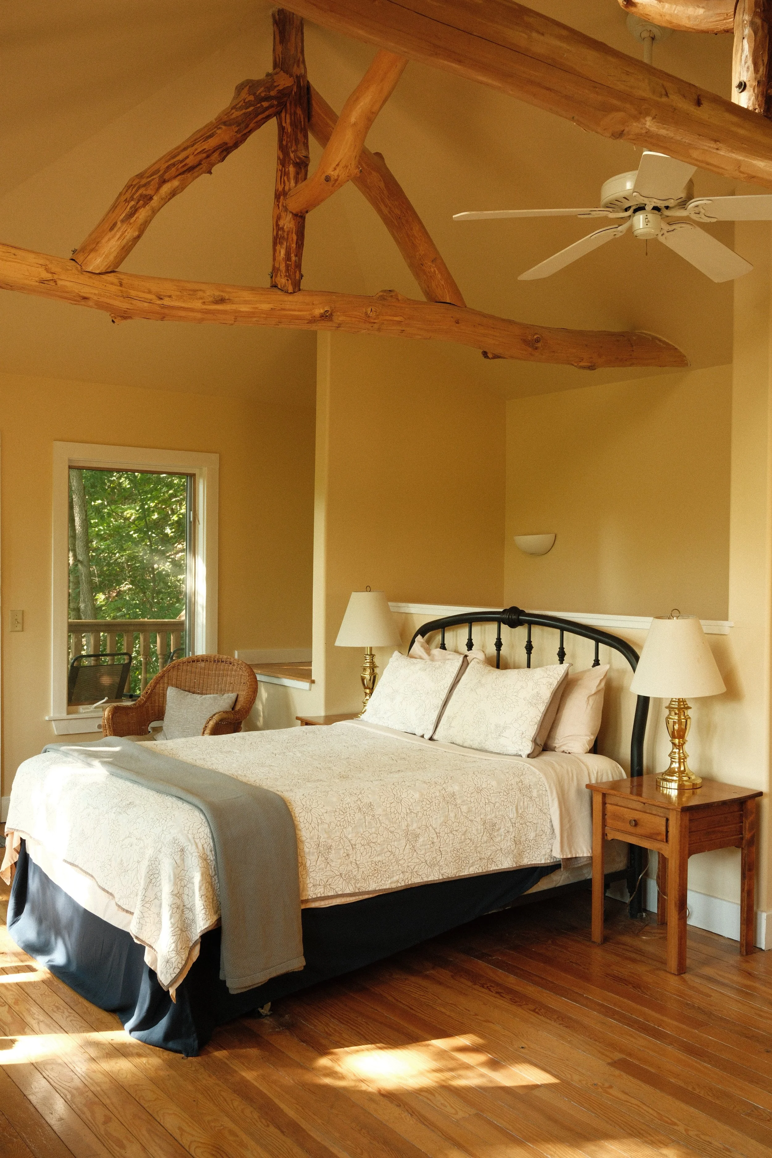 A bedroom with a wooden bed frame, cream bedding, nightstand with a gold lamp, wicker chair, window showing greenery outside, and wooden flooring. The ceiling has exposed wooden beams and a white ceiling fan.