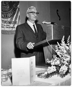 An older man with glasses in a suit standing at a podium with a microphone, next to a bouquet of flowers and a framed document, during a formal event or speech.