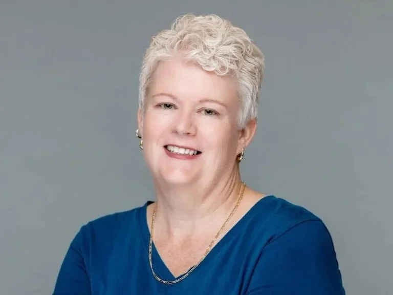 A portrait of a smiling woman with short curly blonde hair, wearing a blue top and gold jewelry, against a plain gray background.