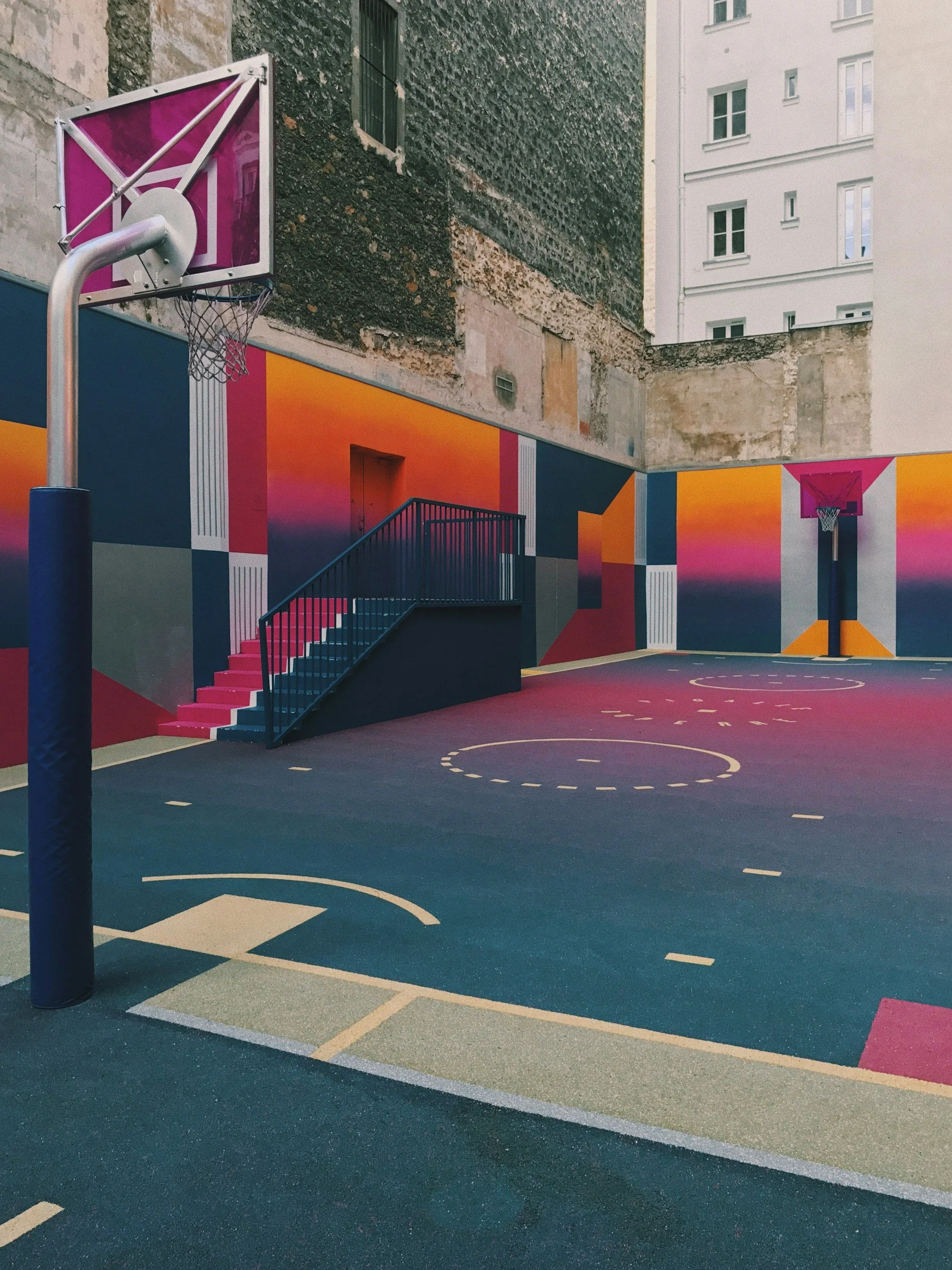 Colorful outdoor basketball court with painted walls featuring geometric patterns, two basketball hoops with pink backboards, and a staircase leading to a door. Urban setting with surrounding buildings.