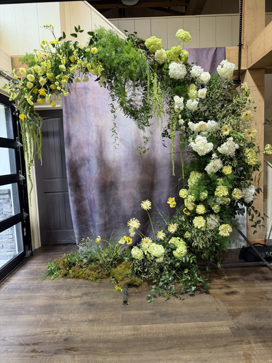 A floral arch made of white and green flowers and foliage, with some yellow accents, set against a backdrop with a grayish misty appearance, on a wooden floor inside a building.