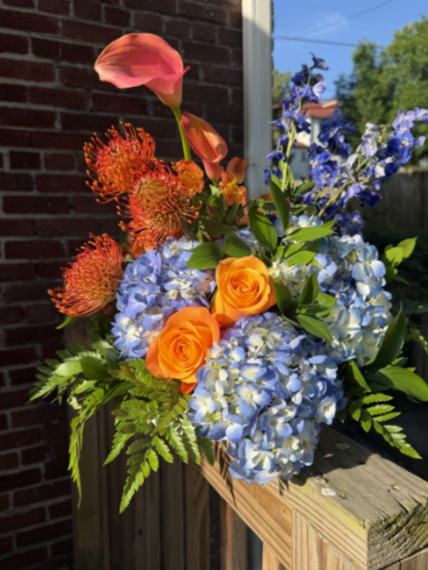 A colorful floral arrangement with pink, orange, and blue flowers, including roses, hydrangeas, and other blooms, sitting on a wooden surface outside near a brick wall and trees.