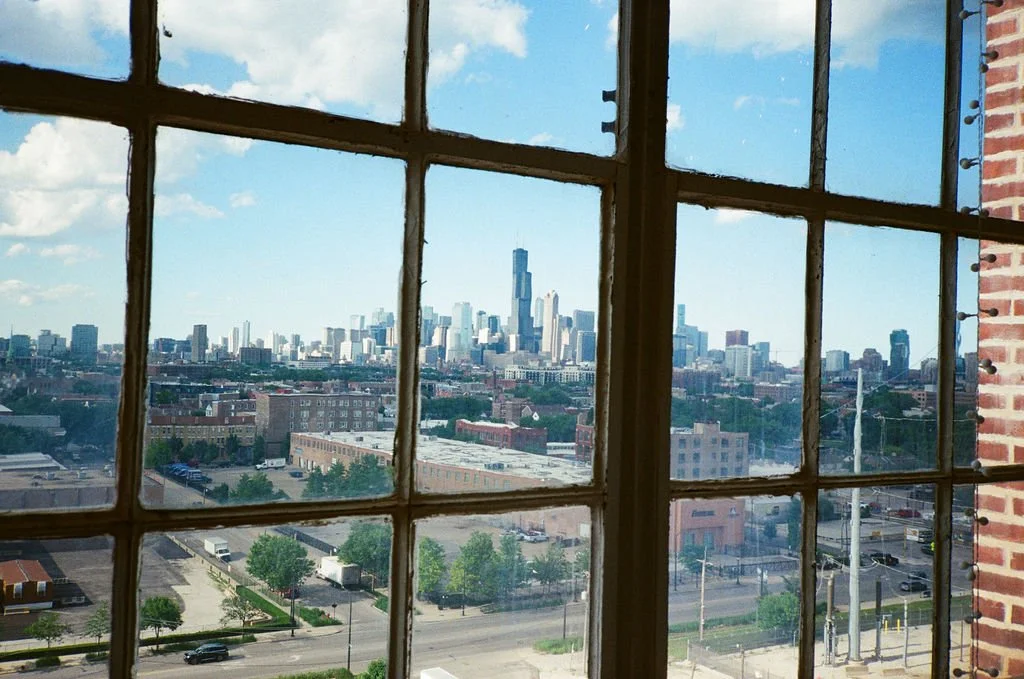 View of a city skyline through a large industrial-style window with grid panes, with a mix of modern and older buildings, including a prominent skyscraper in the distance.