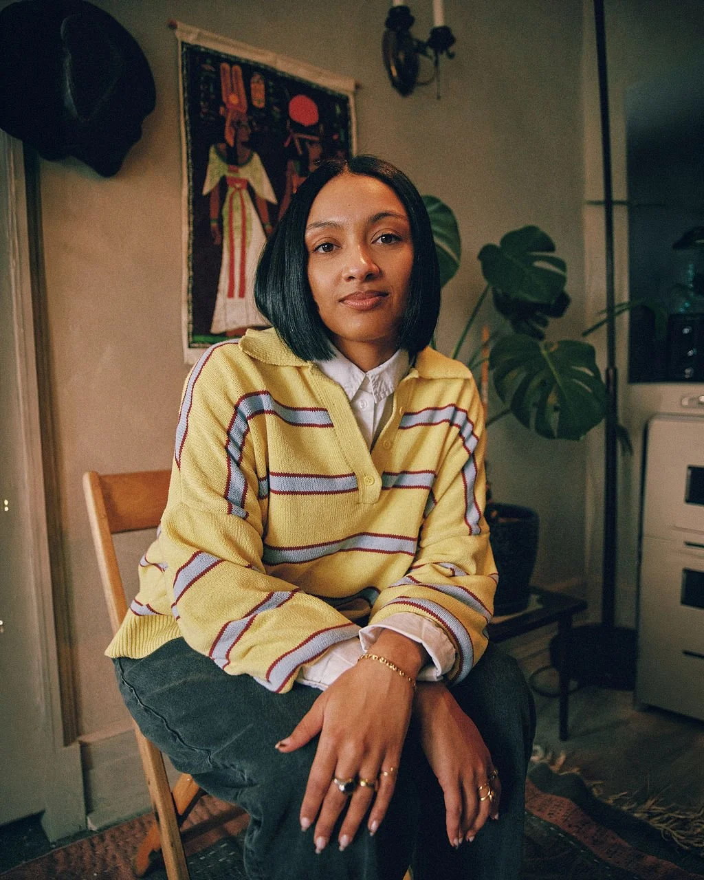 A woman with black hair and medium skin tone sitting on a wooden chair in a cozy room, wearing a striped yellow sweater over a white shirt, with large green plants in the background, and art hanging on the wall.