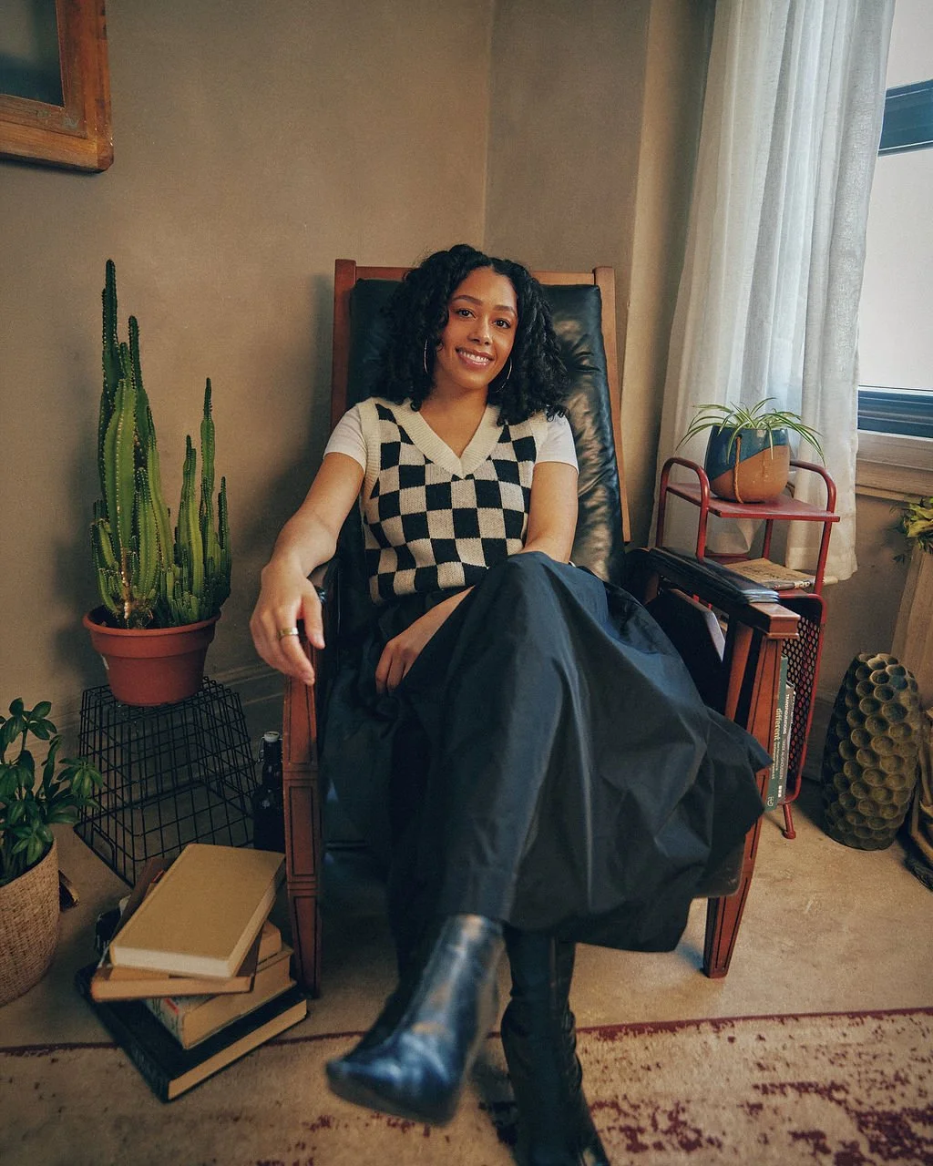 A woman with curly hair sitting in a high-backed chair, smiling, wearing a black and white checkered vest, white t-shirt, black skirt, and black boots, in a cozy room with plants, books, and a window.