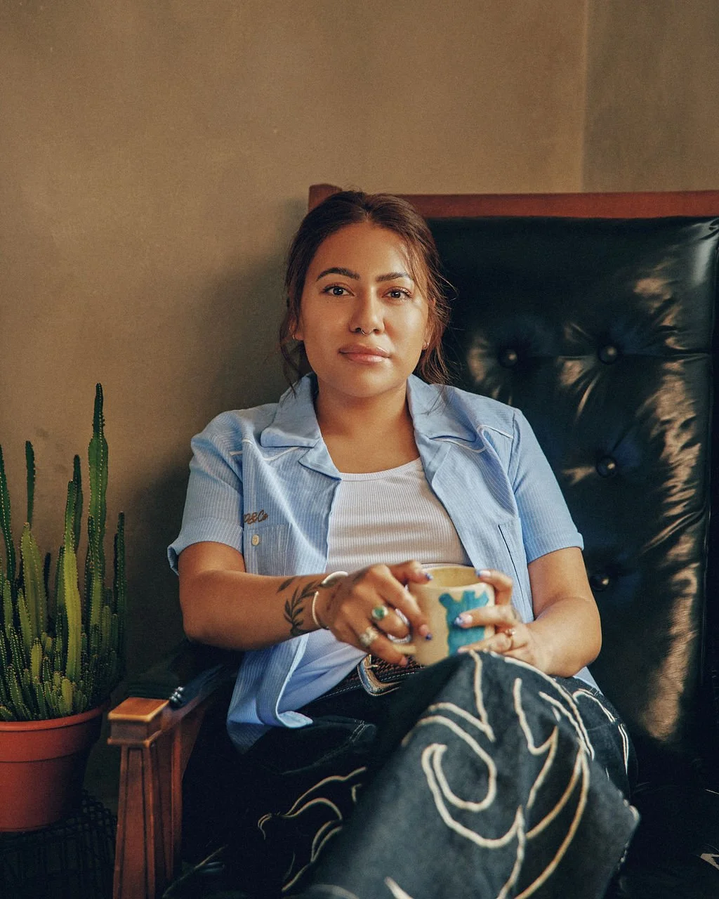 A woman sitting on a black leather armchair, holding a mug, wearing a blue shirt over a white top, with tattoos on her arms, and beside a potted cactus plant.