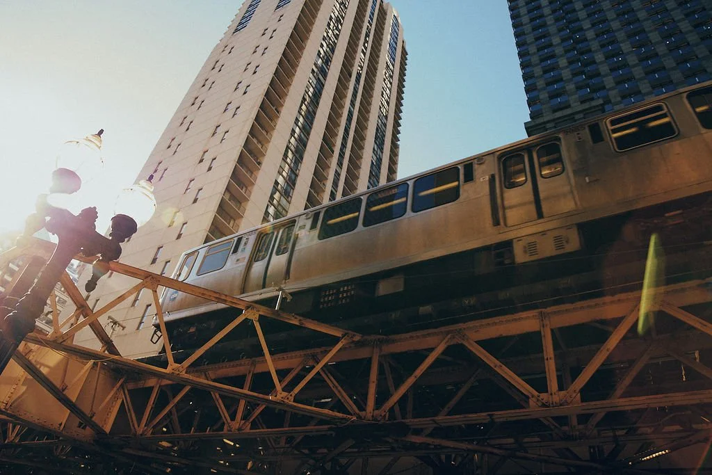 A city scene with a tall building, a train on an elevated track, and a street lamp against a clear sky.