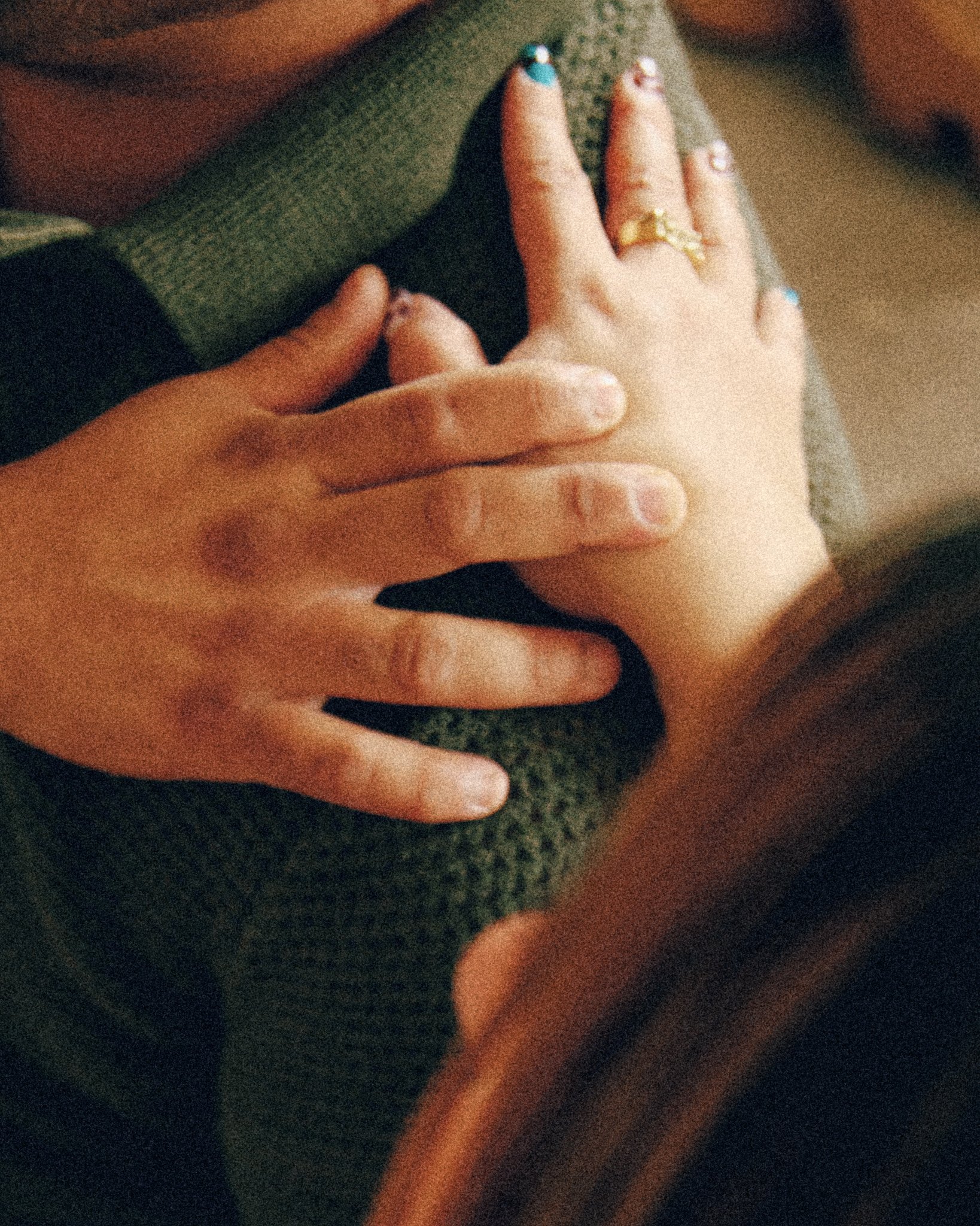 Close-up of a person's hand resting on a person's lap, with painted fingernails and rings, in a dimly lit setting.