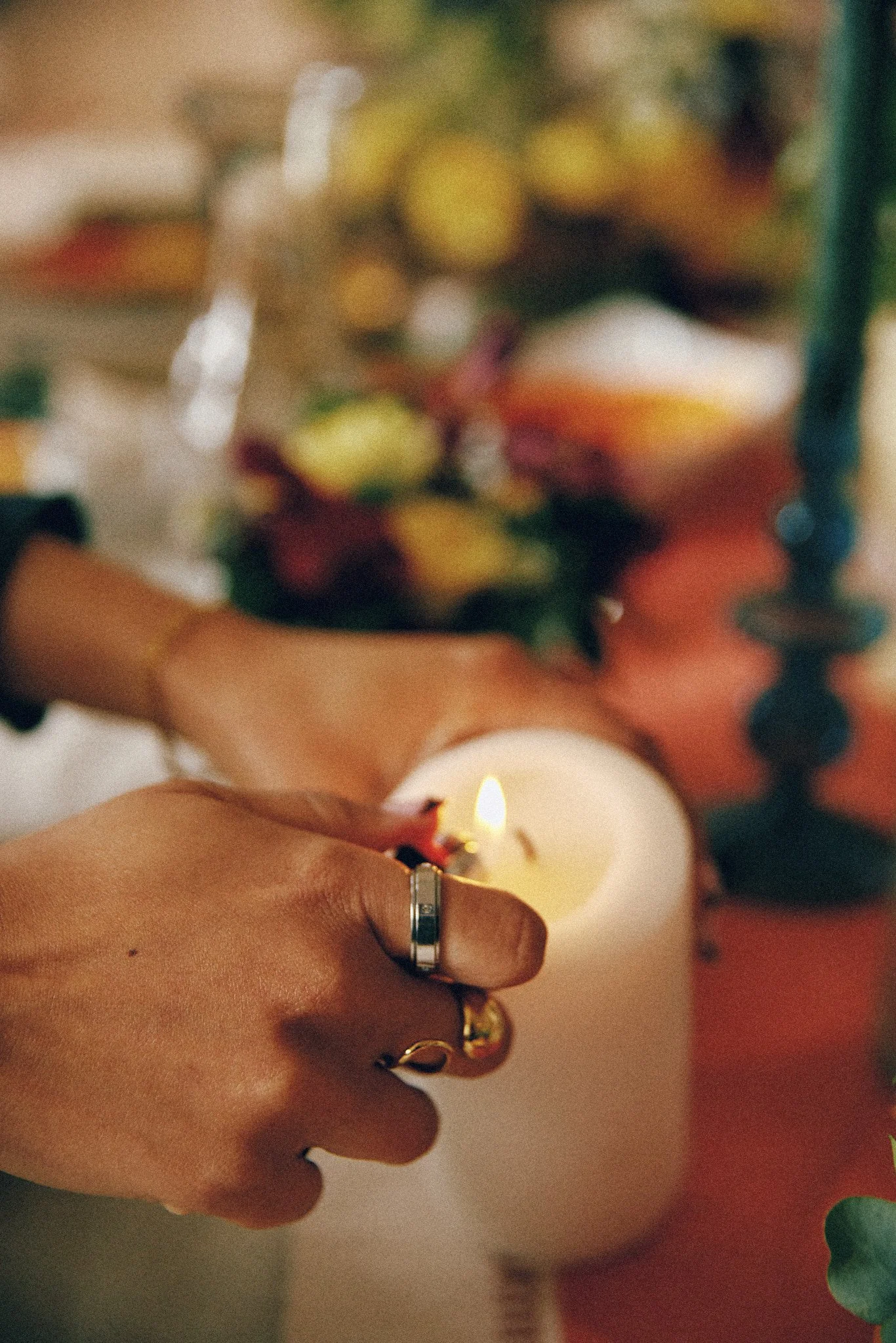 Close-up of a person's hand holding a lit white candle, with blurred colorful floral background.