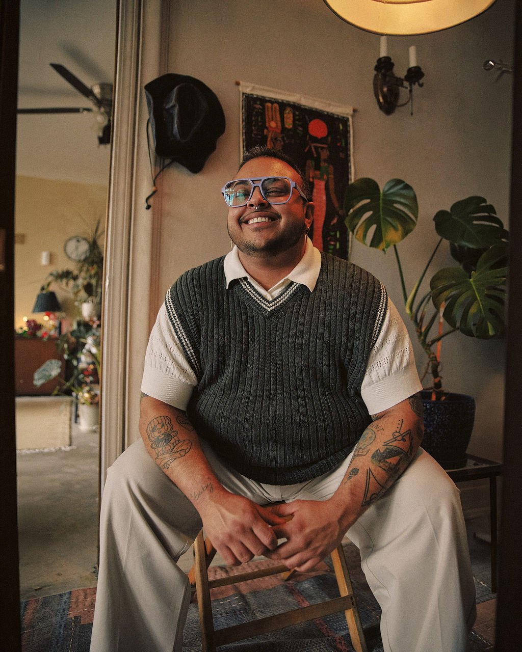 A smiling person sitting on a chair indoors, wearing glasses, a short-sleeved shirt, and a knit vest, with tattoos on their arms, in front of a plant and decorative wall art.