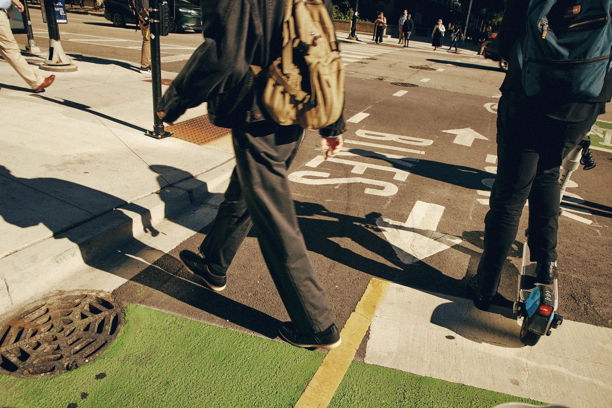 People crossing a city street at a crosswalk with various vehicles parked and moving in the background. The sidewalk features a manhole cover and a section painted green.
