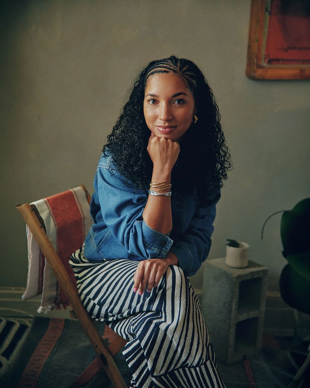 A woman with curly hair and light brown skin, wearing a denim jacket and striped pants, sitting on a wooden chair with her chin resting on her hand, in a cozy room with a beige wall and a small side table.