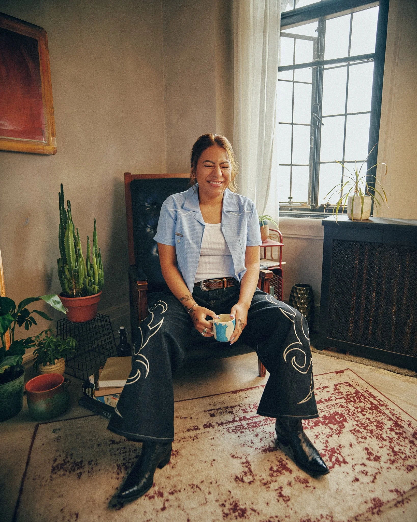 Young woman sitting in a colorful, decorated room with plants and a window, smiling with her eyes closed, holding a mug.