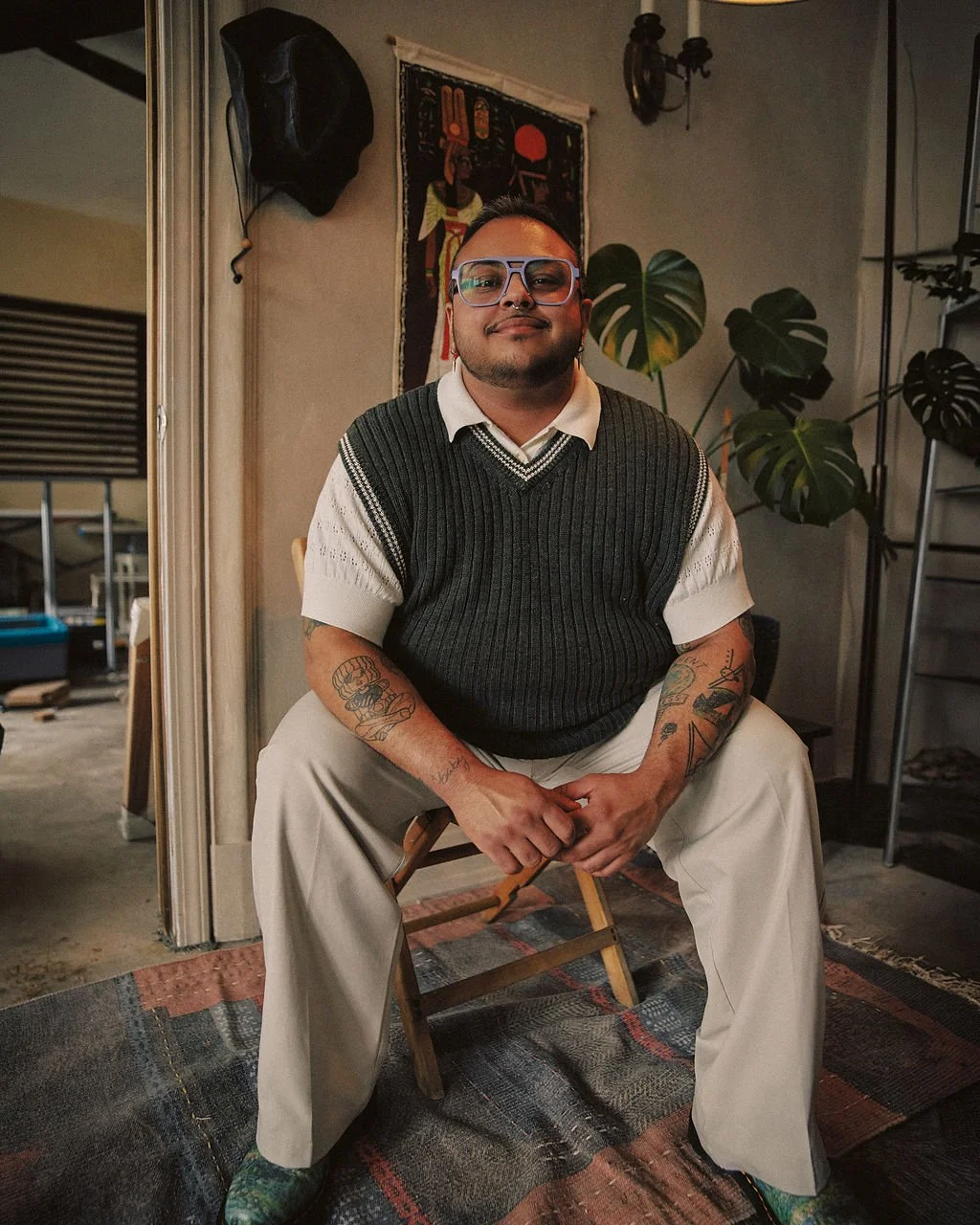 A man with glasses and tattoos sitting on a wooden chair in a room with indoor plants and an art piece on the wall.