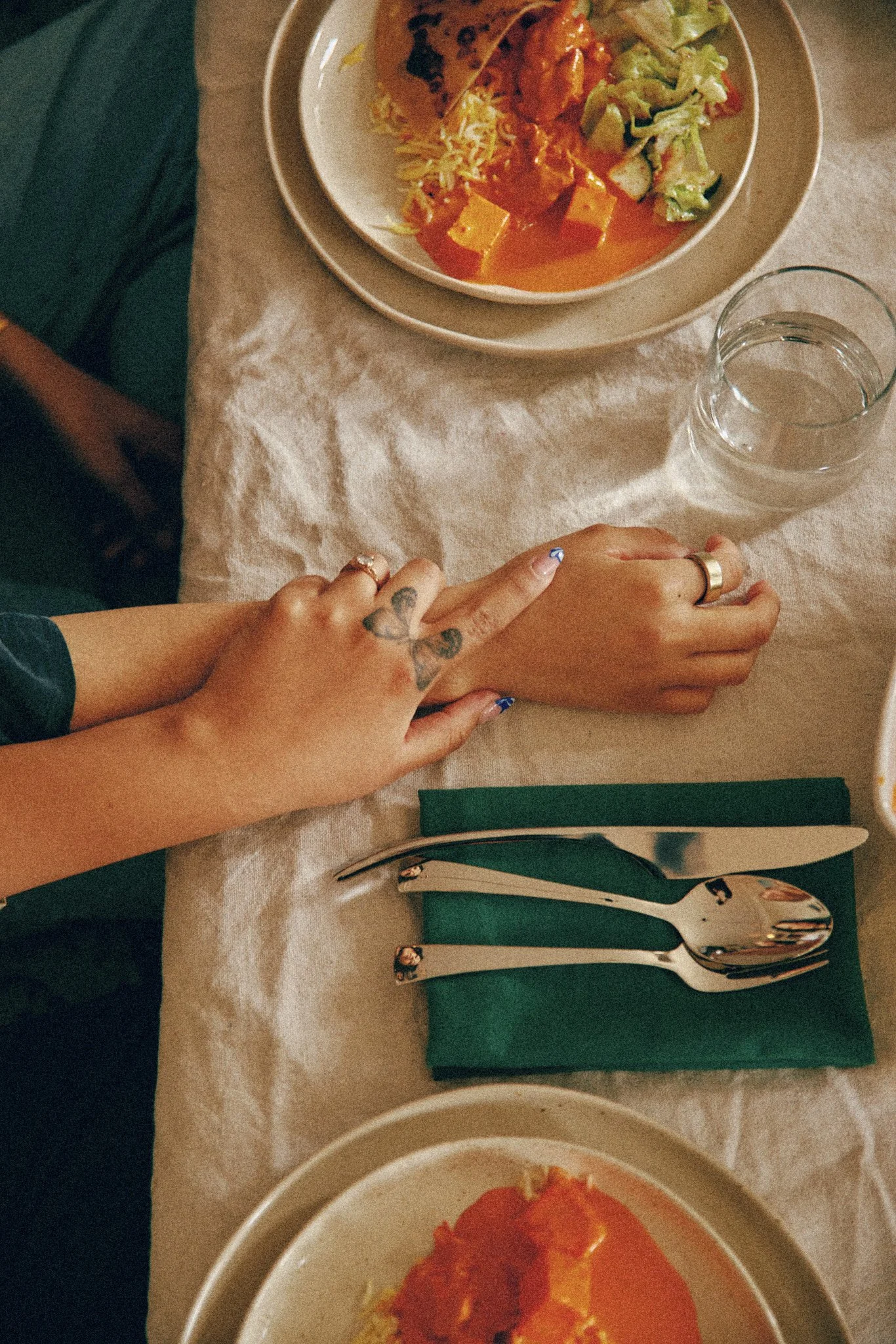 A table with a bowl of food containing rice, a tortilla chip, cheese, lettuce, and a pot of salsa. There is a glass of water, a green folded napkin, and silverware on the table. A person's hands and wrist are visible, with tattoos and rings, holding another person's hand.
