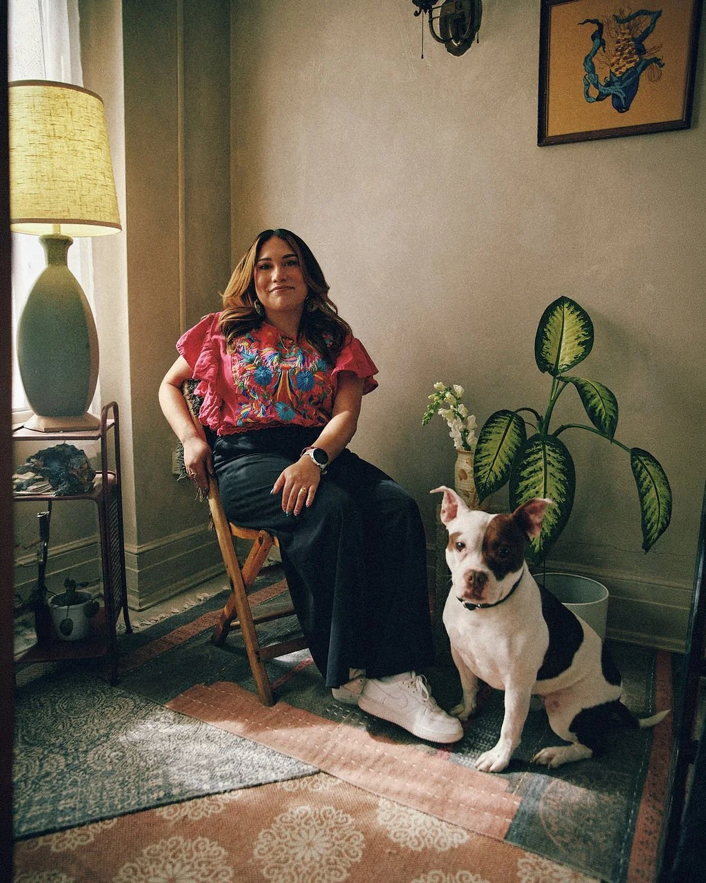 A woman sitting on a wooden chair with a black and white dog nearby in a cozy living room. The woman is wearing a colorful embroidered top and black pants, and is looking at the camera. There is a large potted plant, a lamp, and wall art in the room.