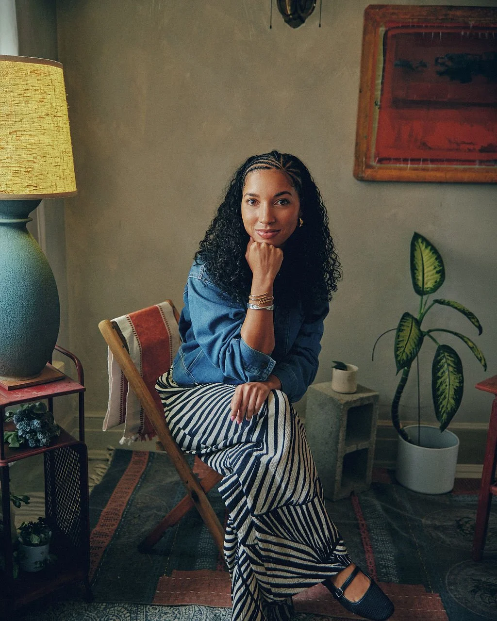 A woman with curly black hair, wearing a denim jacket and striped black and white pants, sitting on a wooden chair in a cozy, decorated room with plants, a lamp, and artwork.