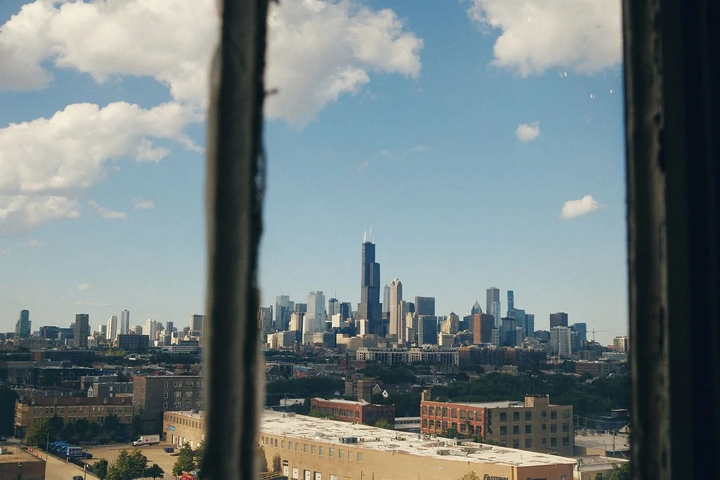 View of the Chicago skyline through a window with bars, featuring the Willis Tower and various skyscrapers against a partly cloudy sky.