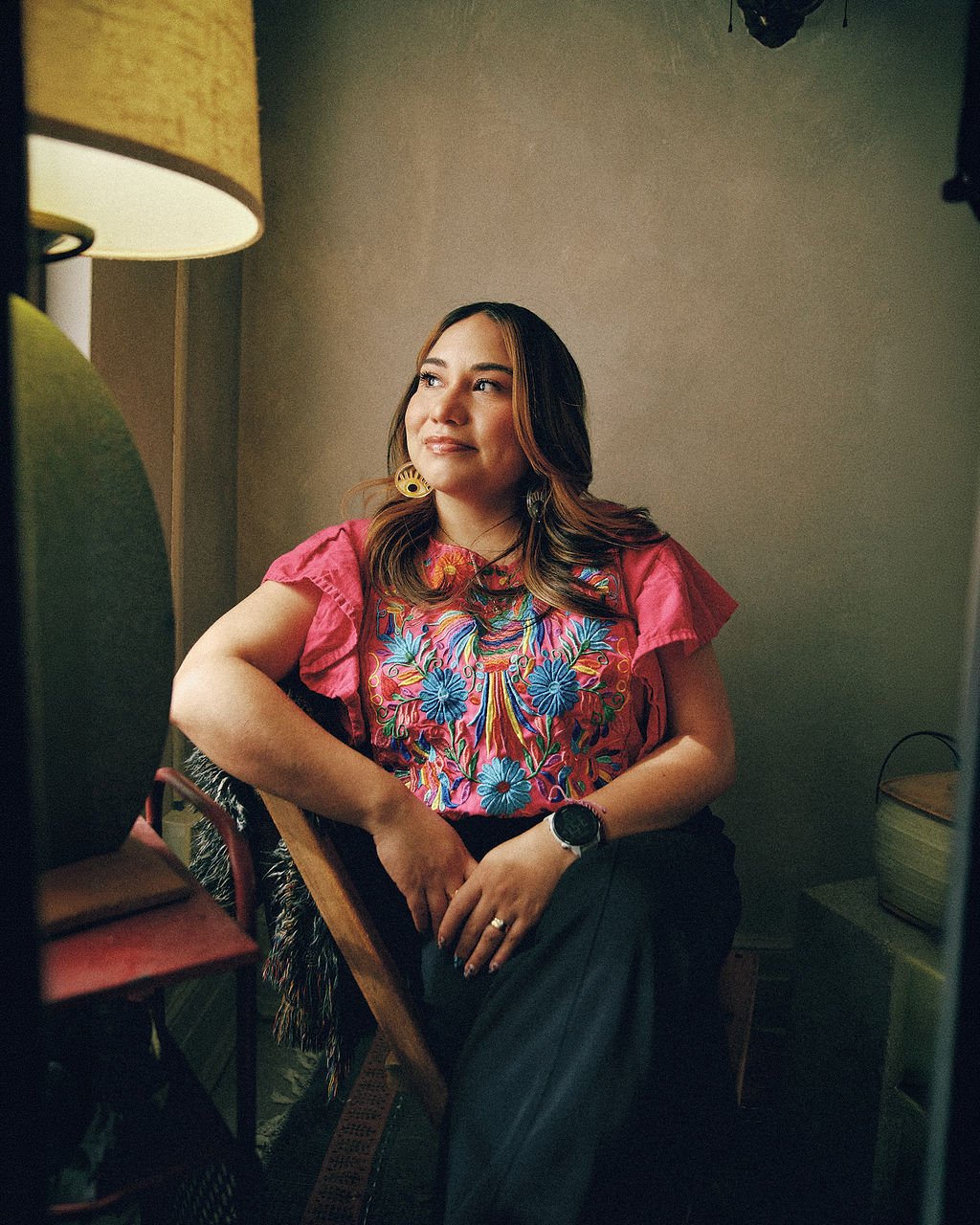 A woman with shoulder-length brown hair sitting on a chair, wearing a colorful floral embroidered top, black pants, gold earrings, and a smartwatch, with soft lighting in the background.